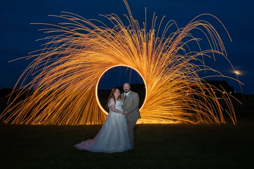 A newlywed couple poses in front of a dramatic long exposure sparkler light painting forming a glowing golden circle against a dark night sky.