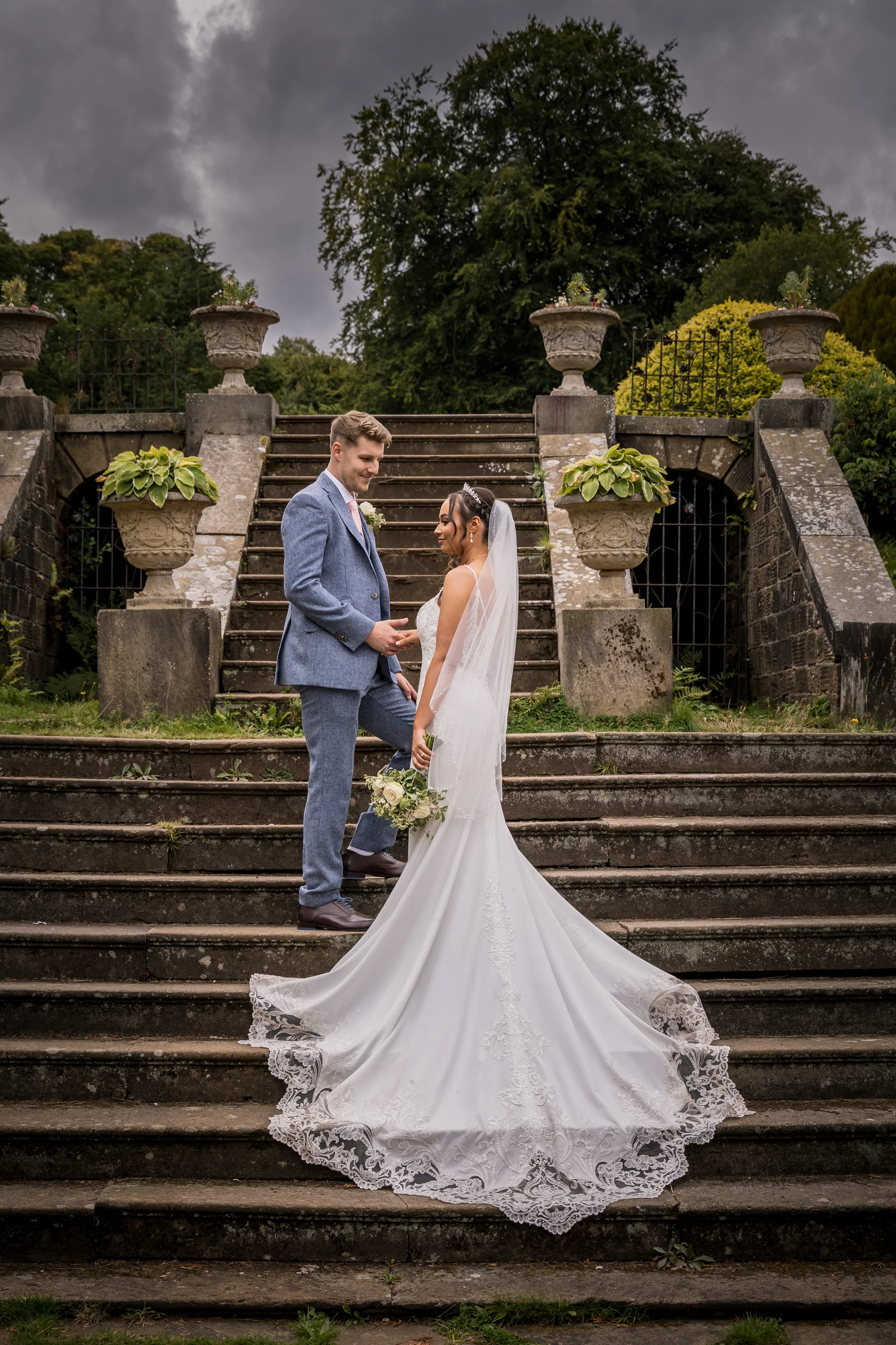 A newlywed couple poses on grand stone outdoor staircase of a country manor under a dramatic cloudy sky, with the bride showcasing a detailed lace train.
