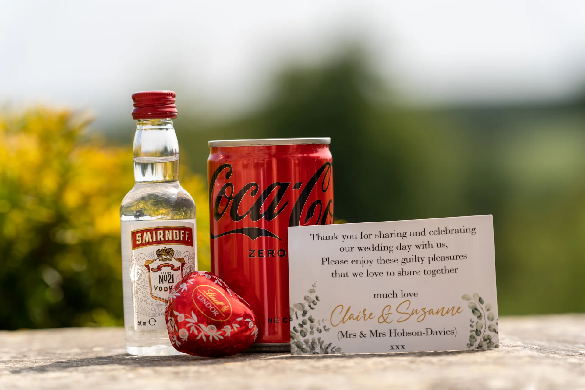 A thoughtful wedding favour set featuring a miniature bottle of vodka, a can of cola, a foil chocolate heart, and a personalised thank you card.