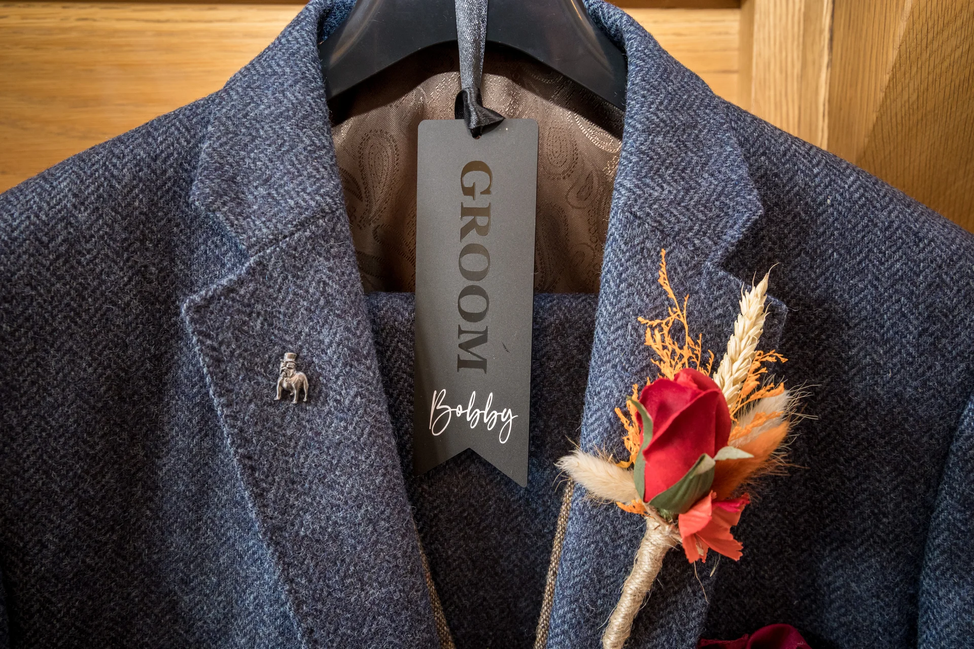 A textured blue tweed suit jacket hangs on a hanger, featuring a personalized groom tag, a silver lapel pin, and a vibrant boutonniere with a red rose and dried florals.