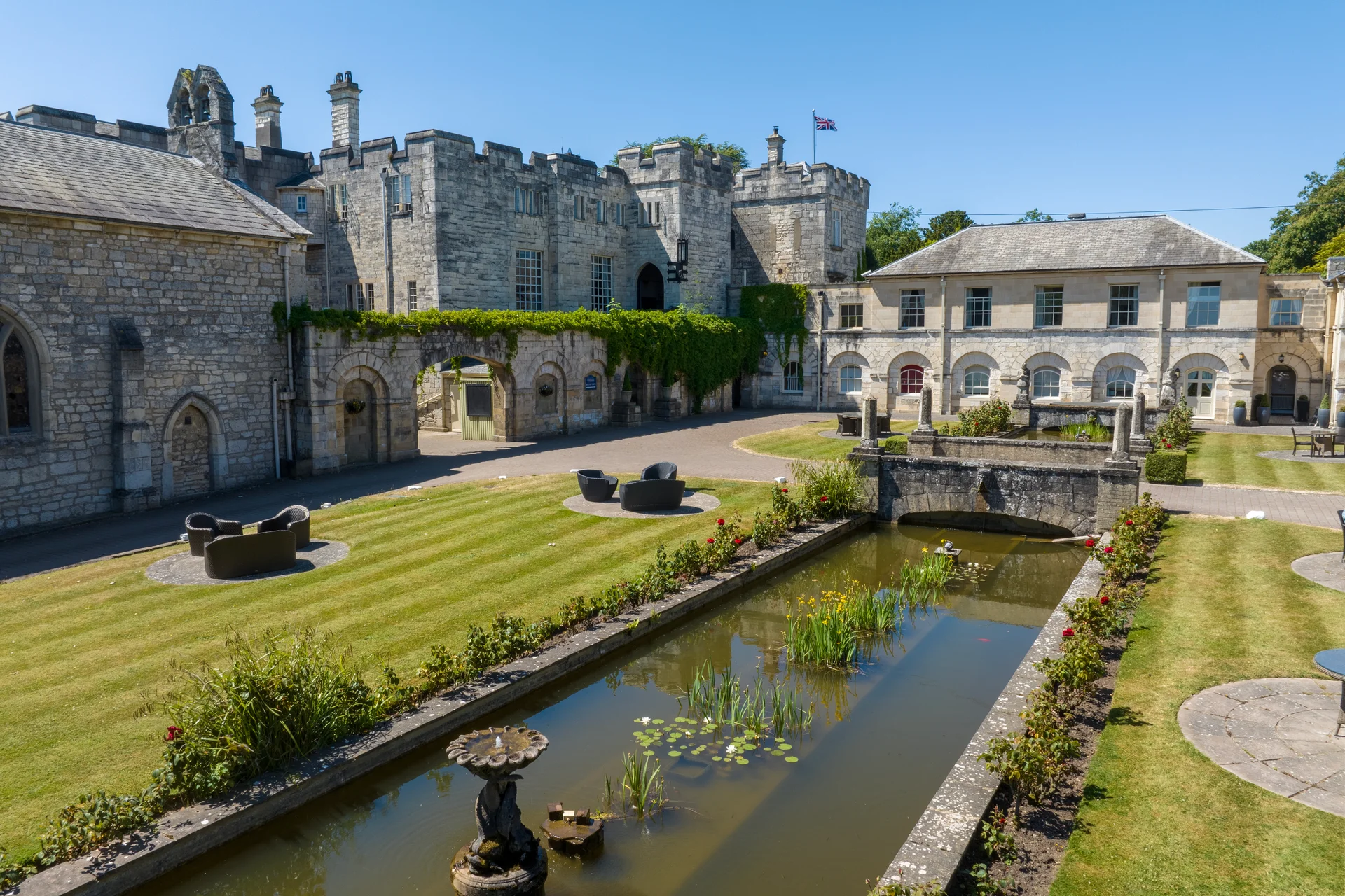 An elevated view of a historic stone castle and country house venue featuring manicured lawns, a long ornamental reflecting pool, stone bridge, and clear blue skies.