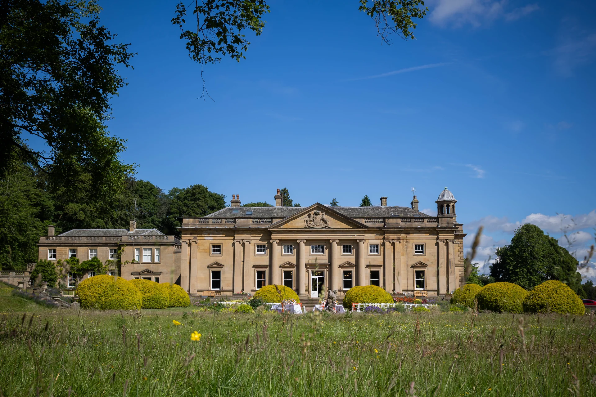 A grand stately home wedding venue sits bathed in golden summer sunshine behind a lush wildflower meadow under a brilliant blue sky.