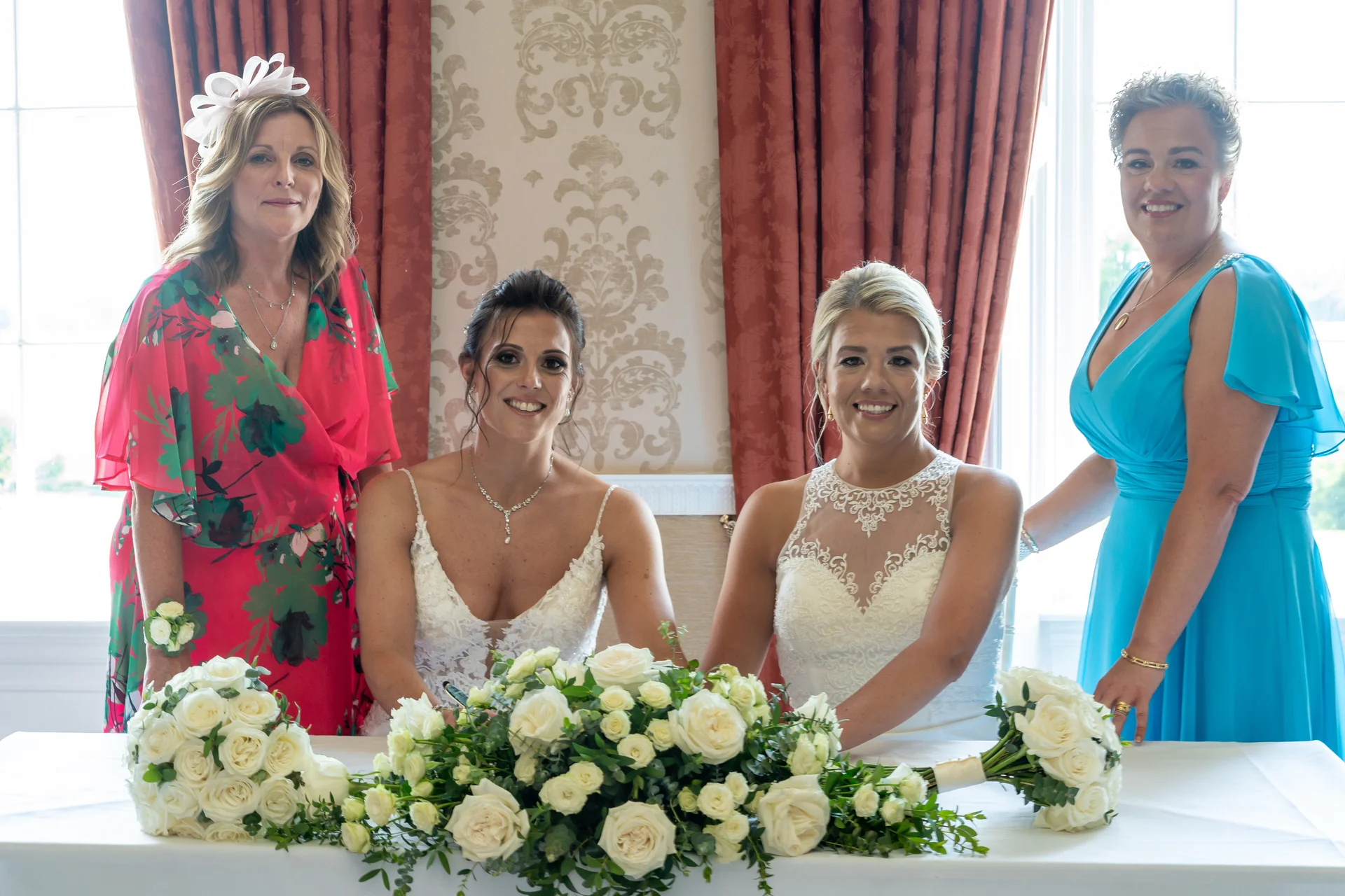 Two brides smile while signing their marriage register, accompanied by family members in brightly coloured dresses and surrounded by white rose floral arrangements.