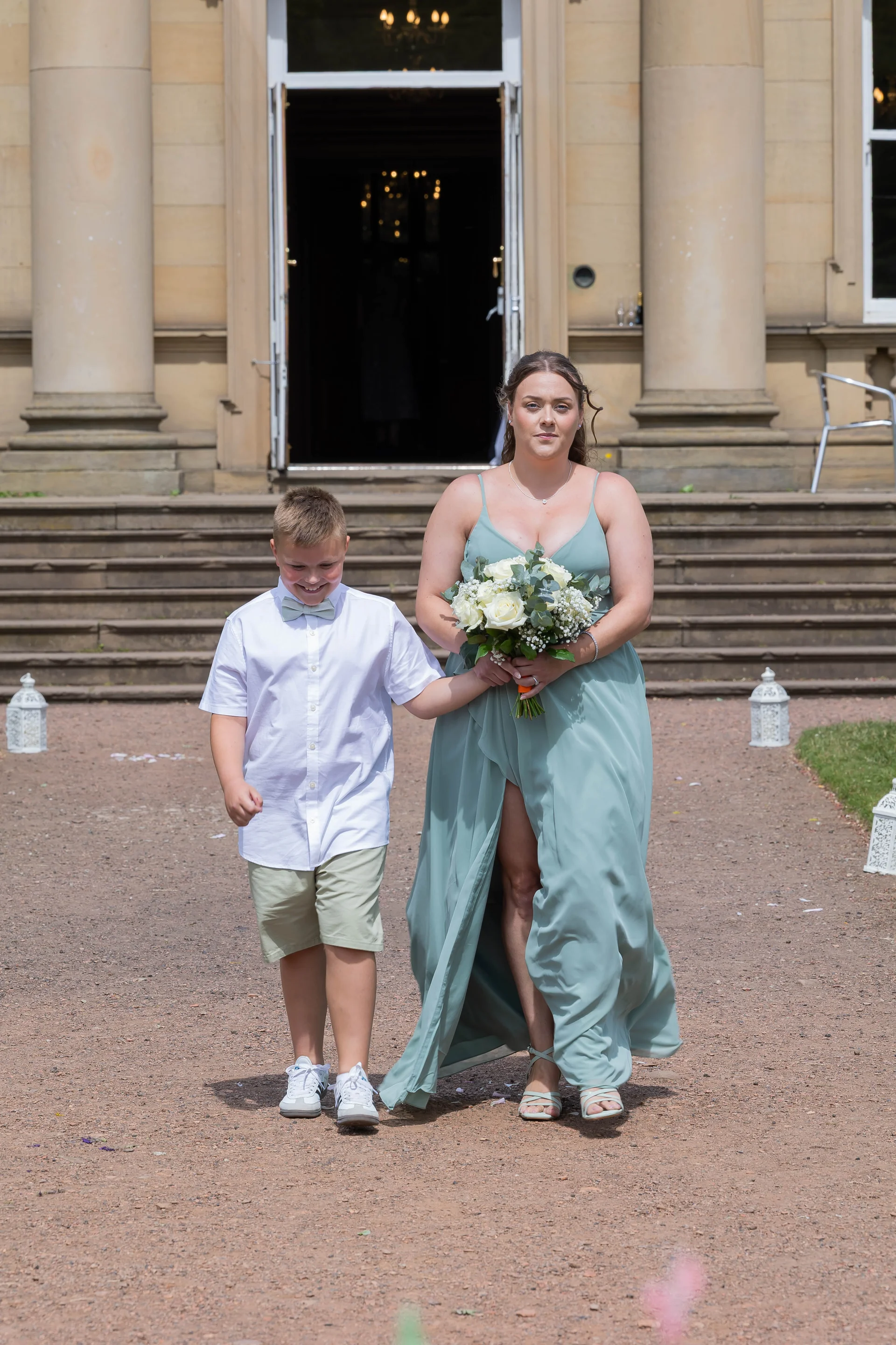 A page boy and a bridesmaid in a sage green dress walk hand in hand down a gravel path outside a grand stately home entrance.