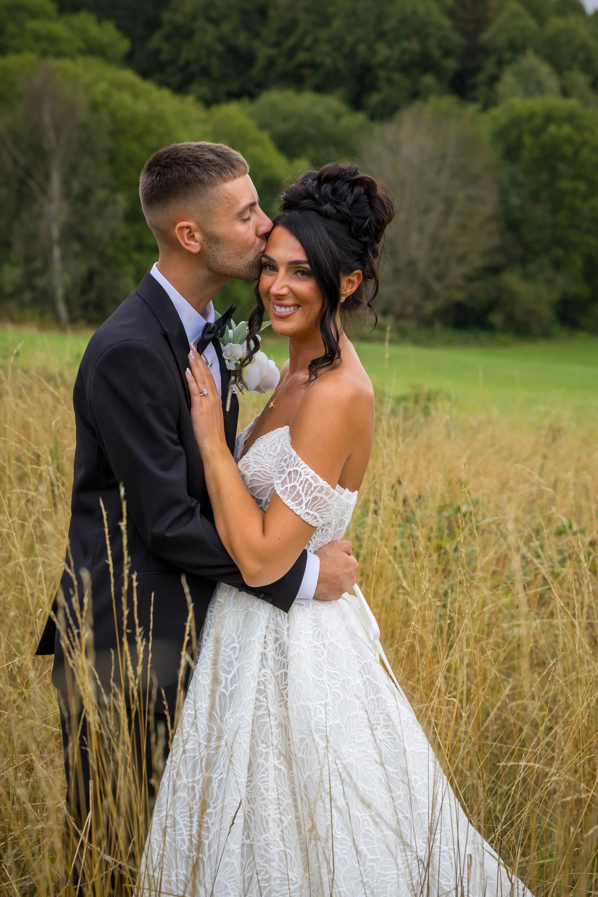 A groom gently kisses his smiling bride on the forehead as they embrace standing amidst tall grass with a rolling woodland backdrop.