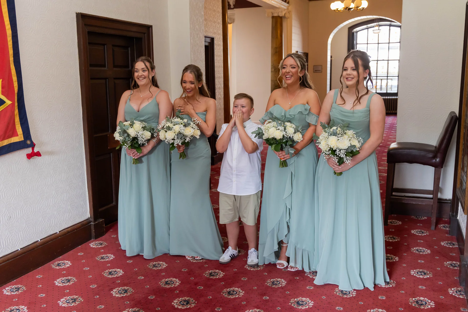 Four bridesmaids in sage green dresses and a page boy react with joyful laughter while holding bouquets of white roses and baby's breath indoors.