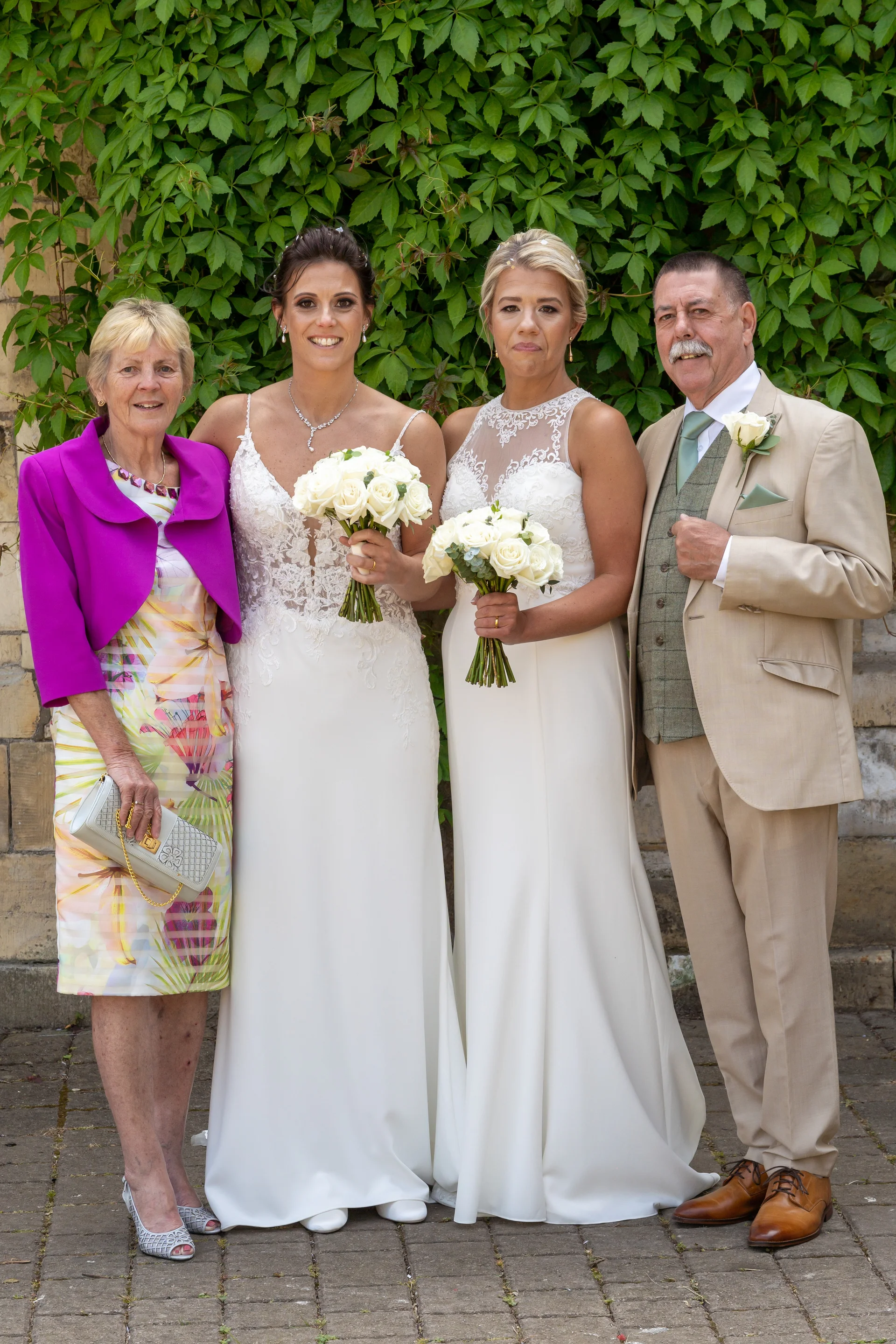 A same-sex couple in elegant white wedding gowns stand alongside family members holding white rose bouquets in front of an ivy-covered stone wall.