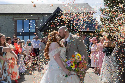 Newlyweds share a romantic kiss outside a stone barn venue as wedding guests shower them with a vibrant cascade of multi-coloured confetti.