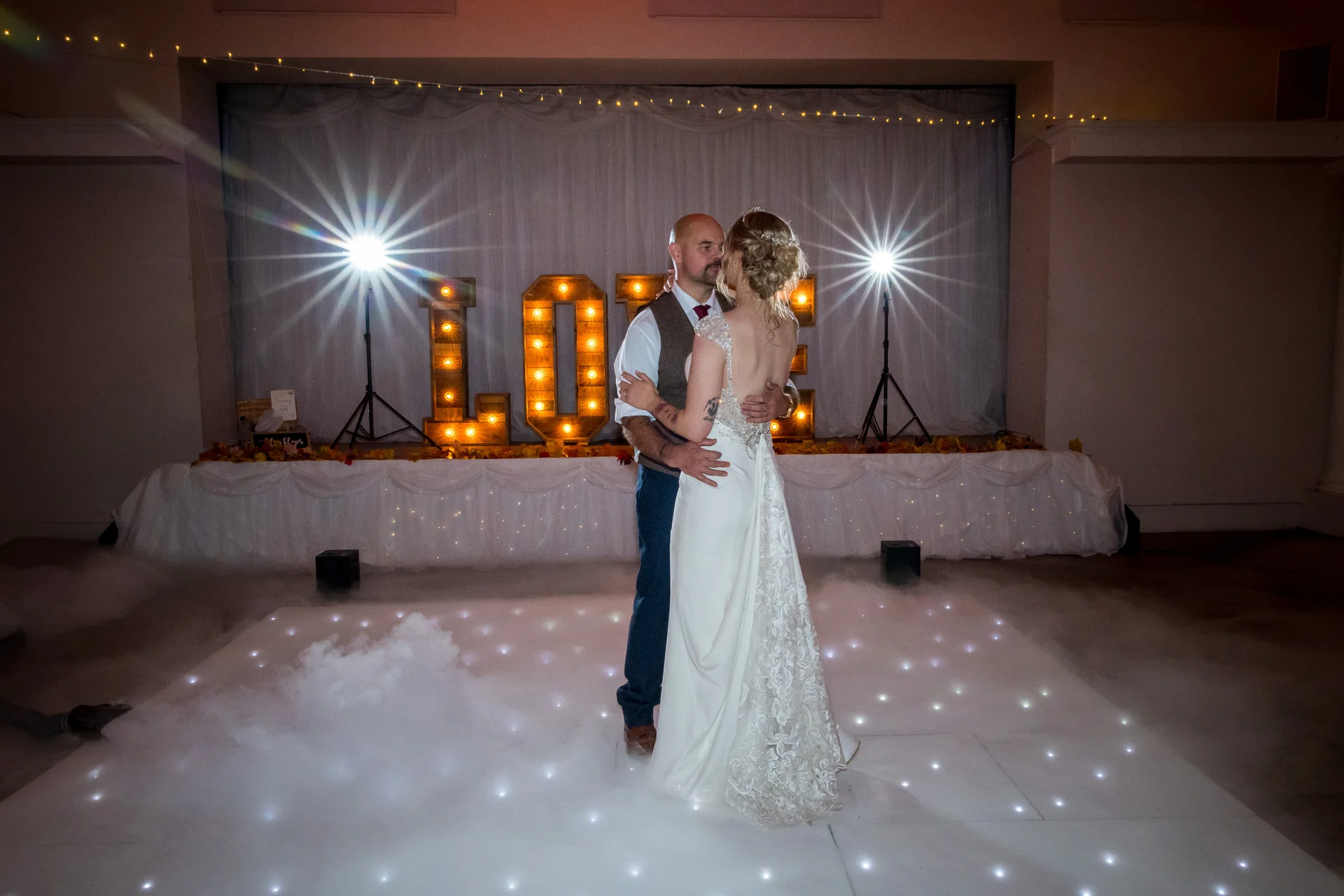 Newlyweds share an intimate moment during their first dance on a lit dance floor surrounded by low-lying fog and glowing letters.