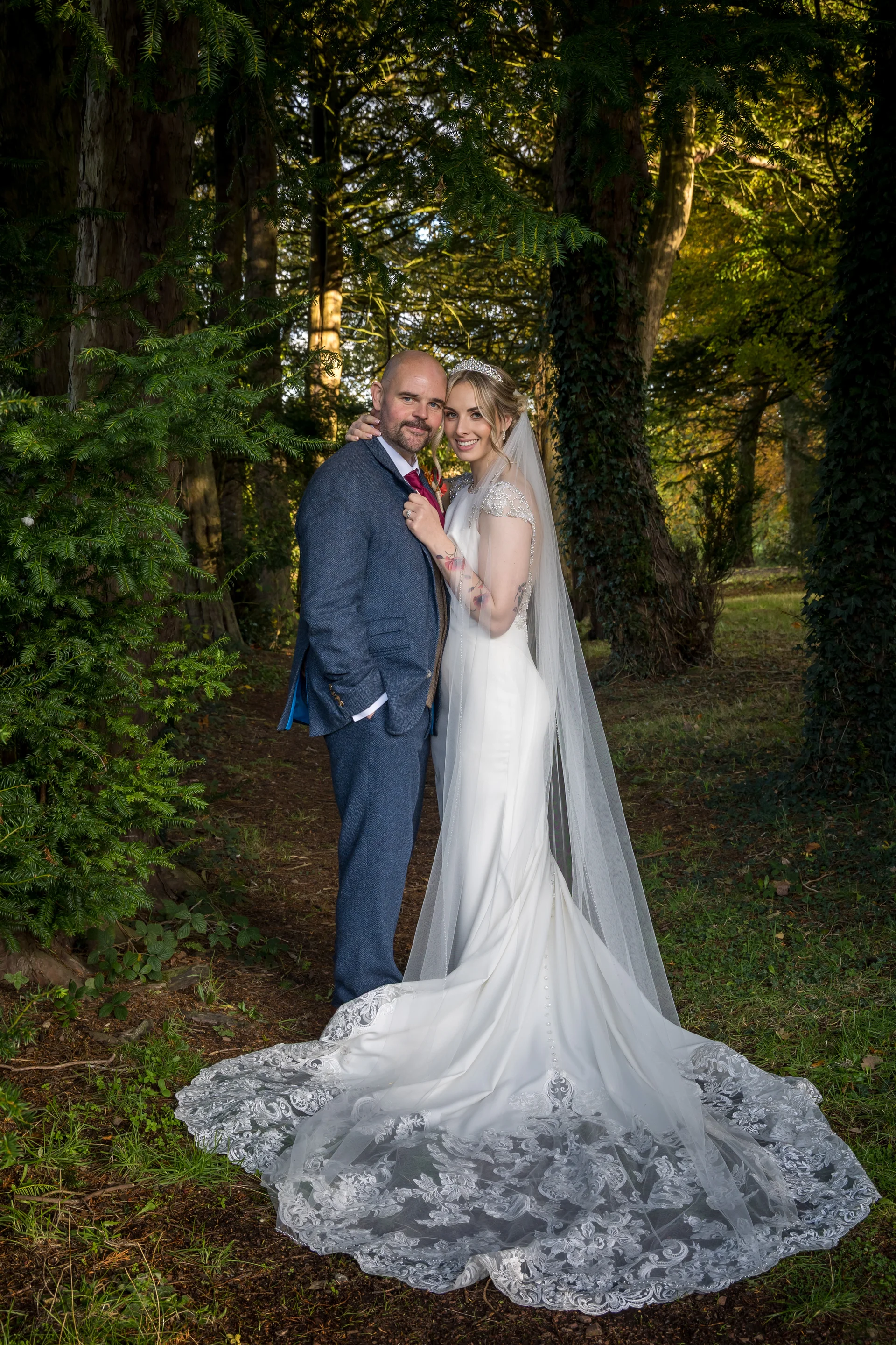 A newlywed couple poses in a sunlit woodland setting, with the bride wearing a sleek gown and long veil, and the groom in a blue tweed suit.