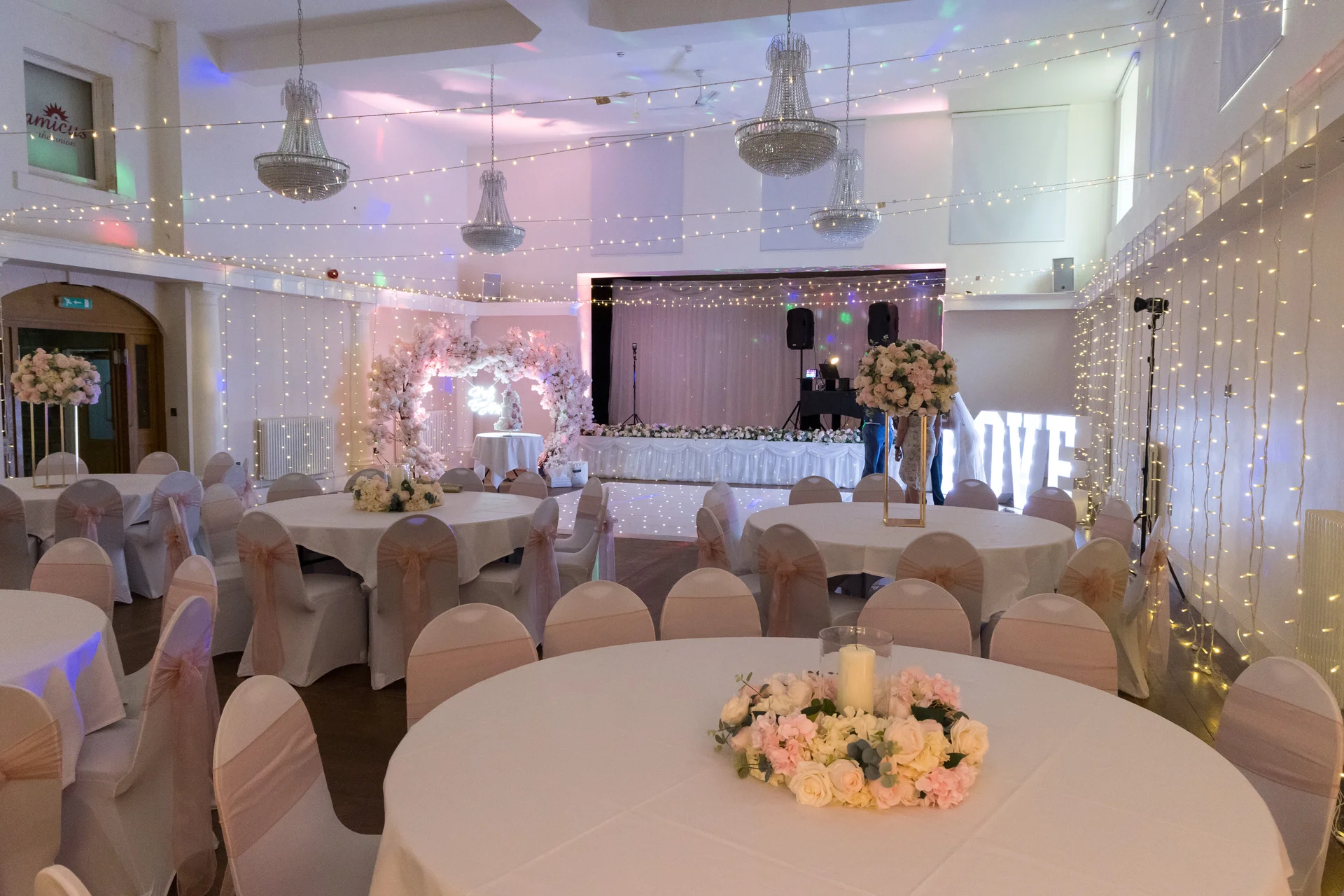 A wedding reception hall is warmly lit with fairy light canopies, crystal chandeliers, round tables featuring blush floral centerpieces, and a glowing light-up love sign at the back of the room.
