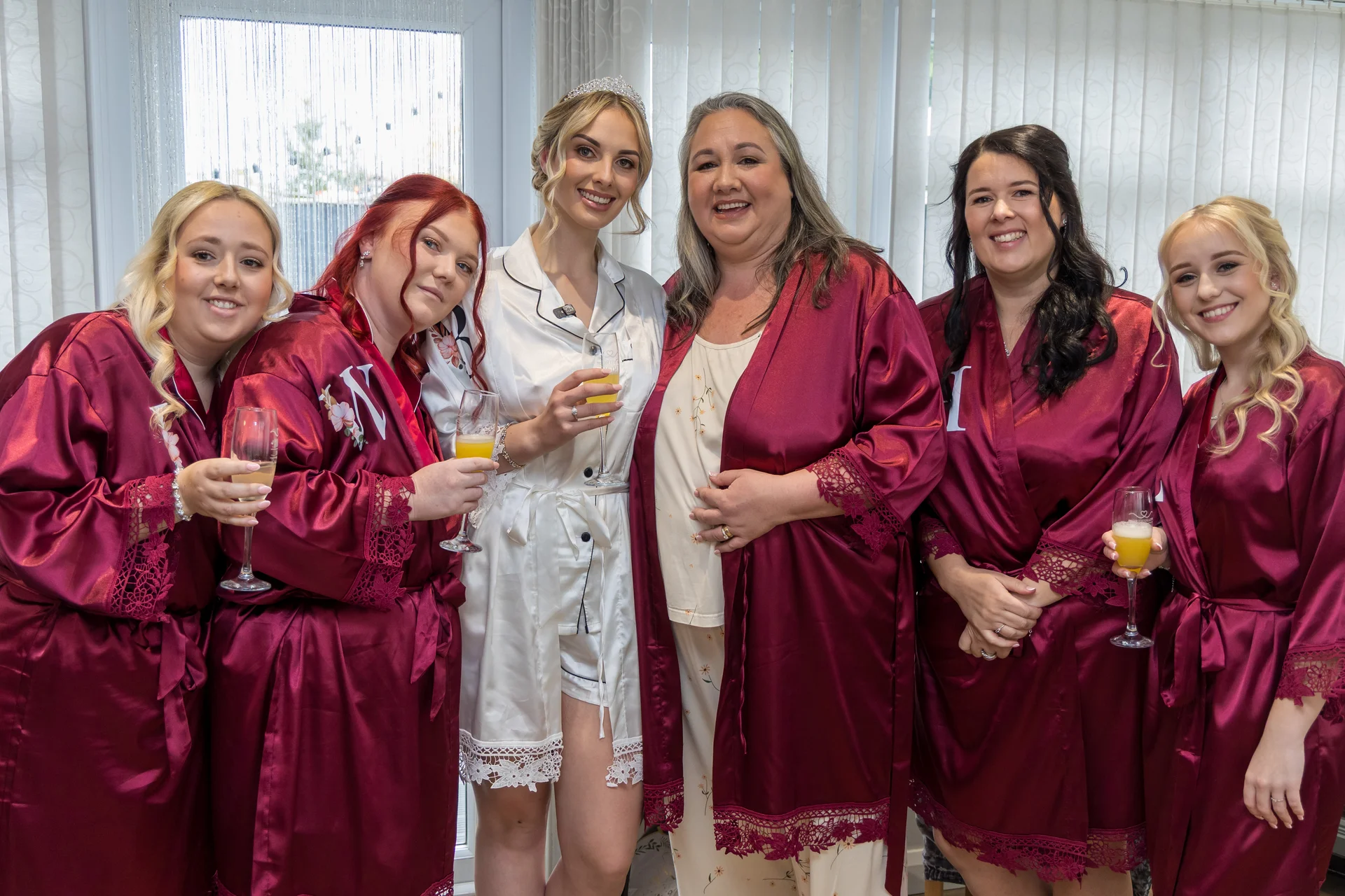 A smiling bride wearing a white satin robe and tiara poses with her bridesmaids and family members who wear matching burgundy satin robes while holding champagne flutes during bridal morning preparations.