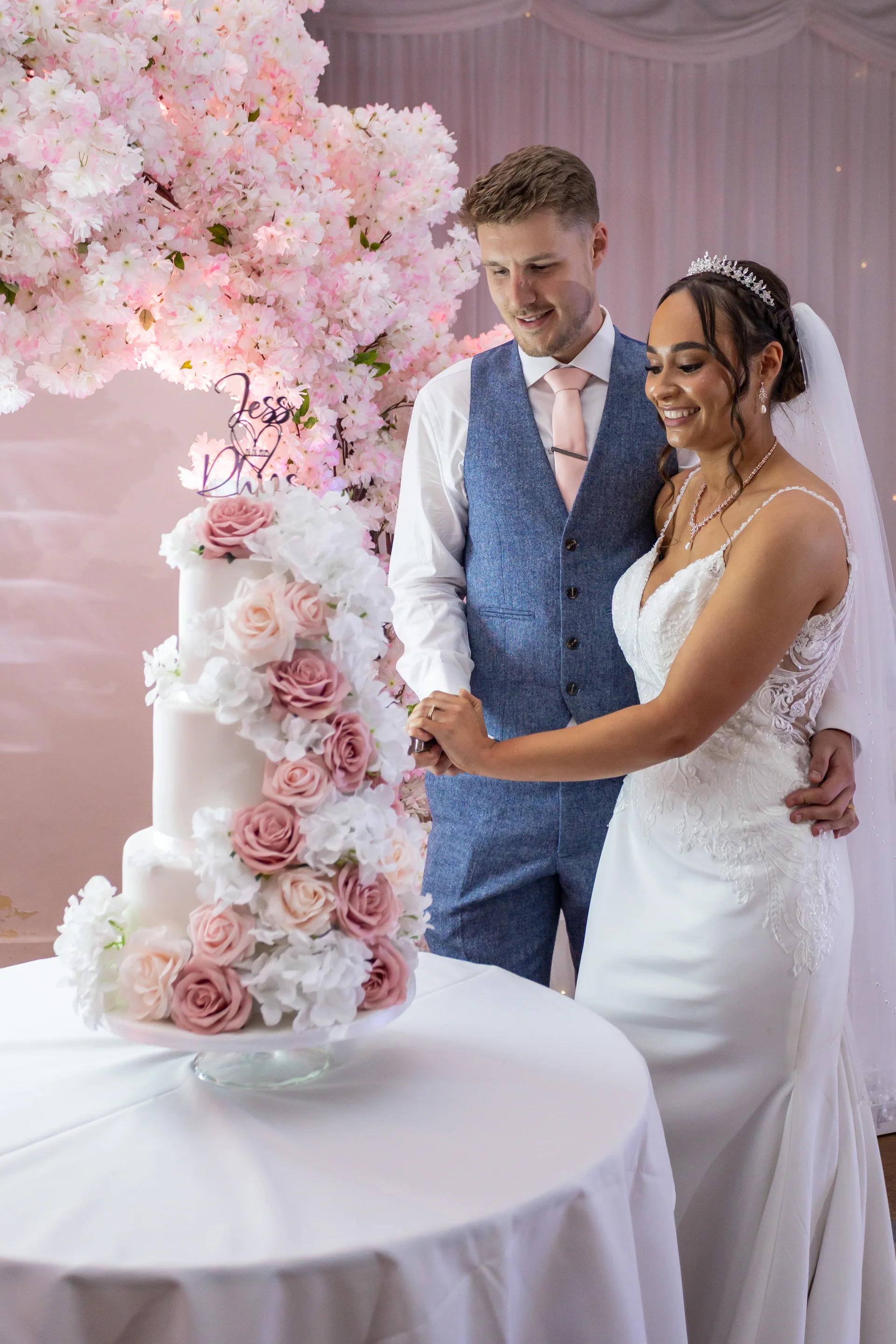 A smiling bride in a lace gown and tiara cuts a three-tier floral wedding cake alongside her groom in a tweed waistcoat beneath a pink blossom arch.