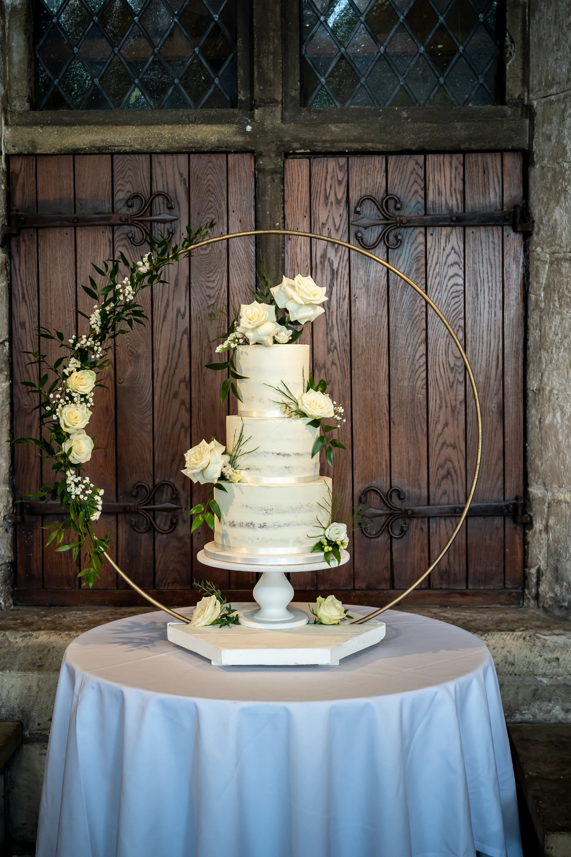 A three-tier semi-naked wedding cake adorned with white roses is displayed on a white cake stand before a gold hoop backdrop and rustic wooden doors.
