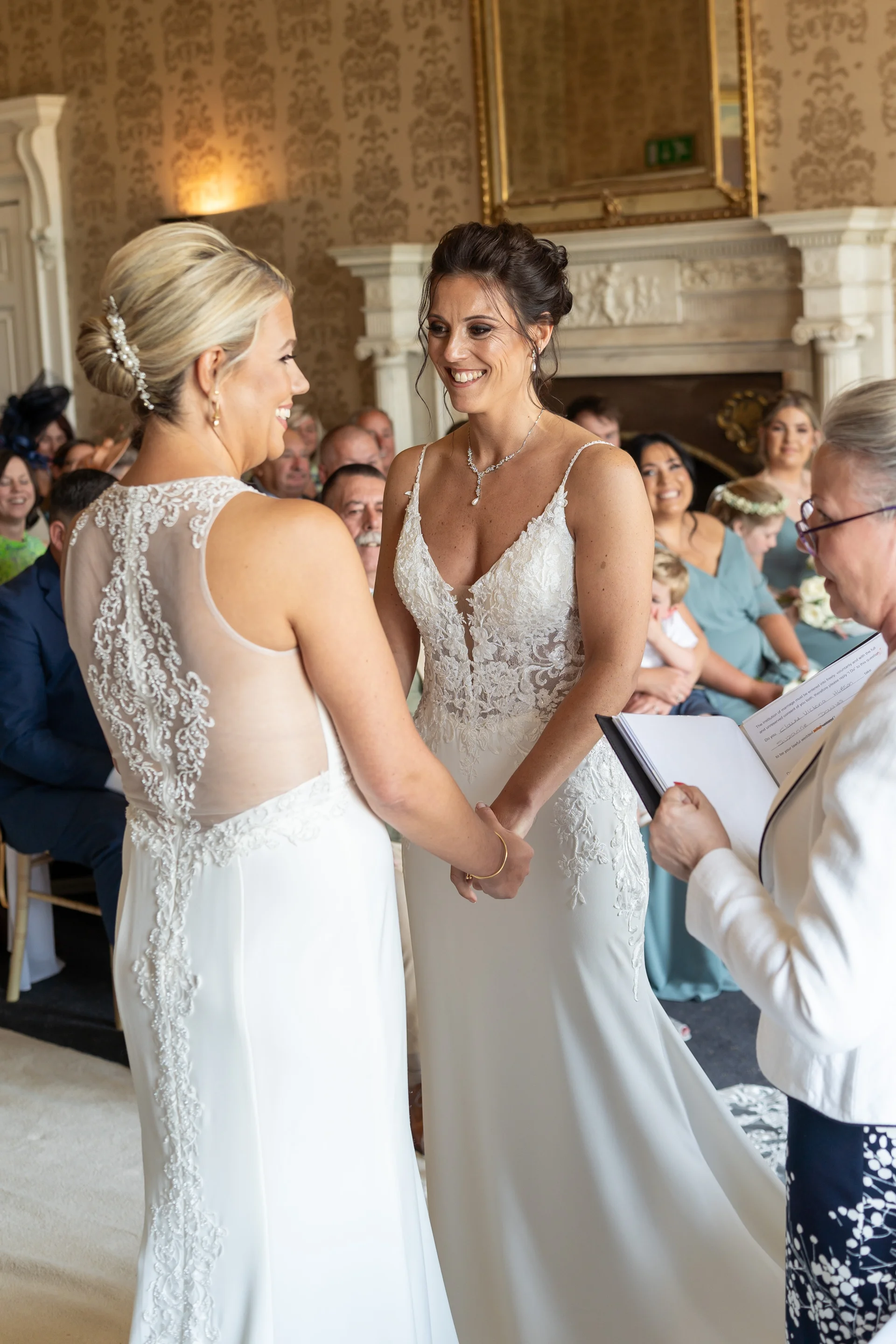 Two brides smiling lovingly at each other while holding hands during their wedding ceremony officiated by a celebrant in a historic manor house room.