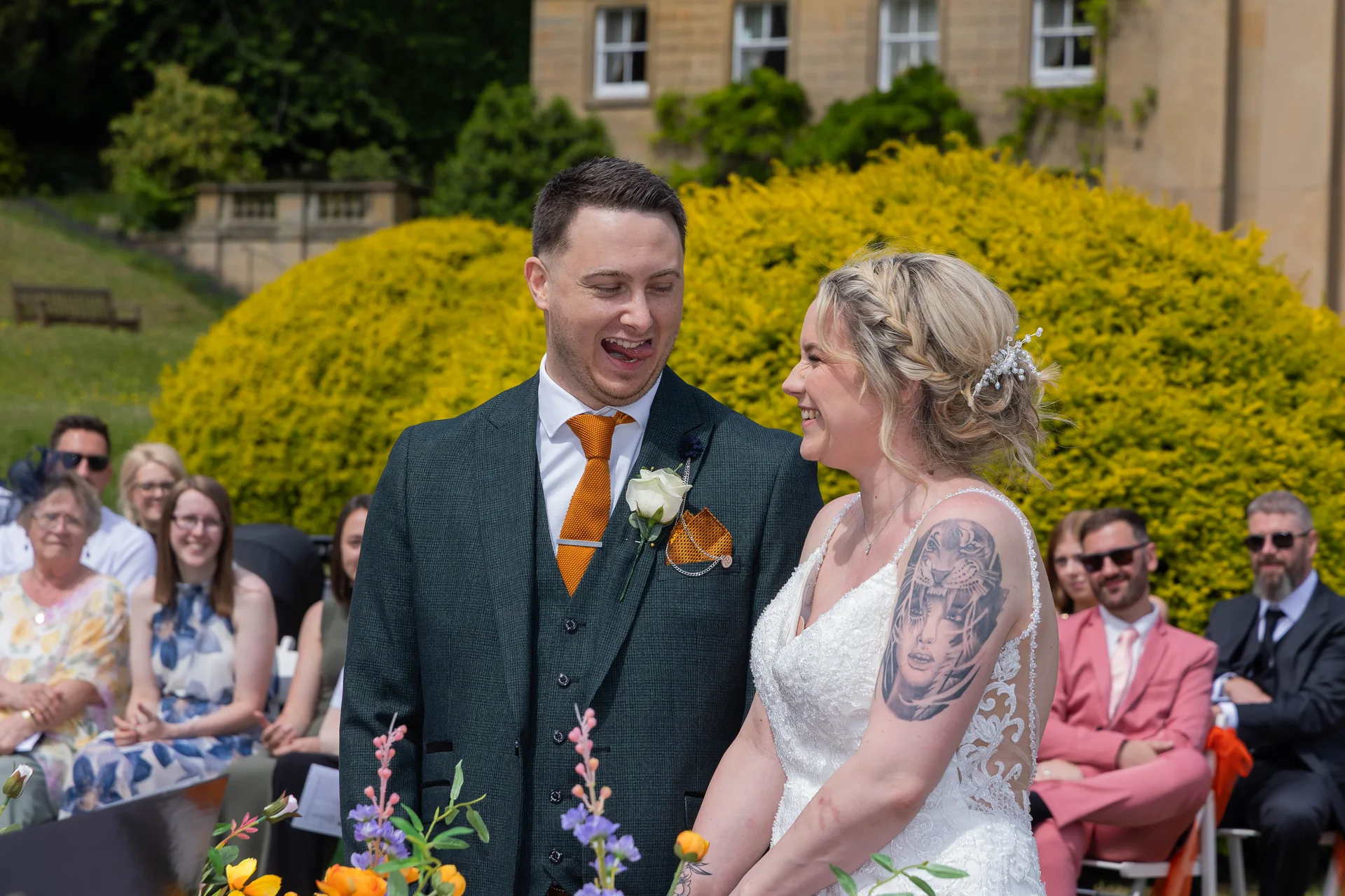 A bride with a detailed arm tattoo and a groom in a dark green three-piece suit share a playful, laughing moment during an outdoor country house ceremony.