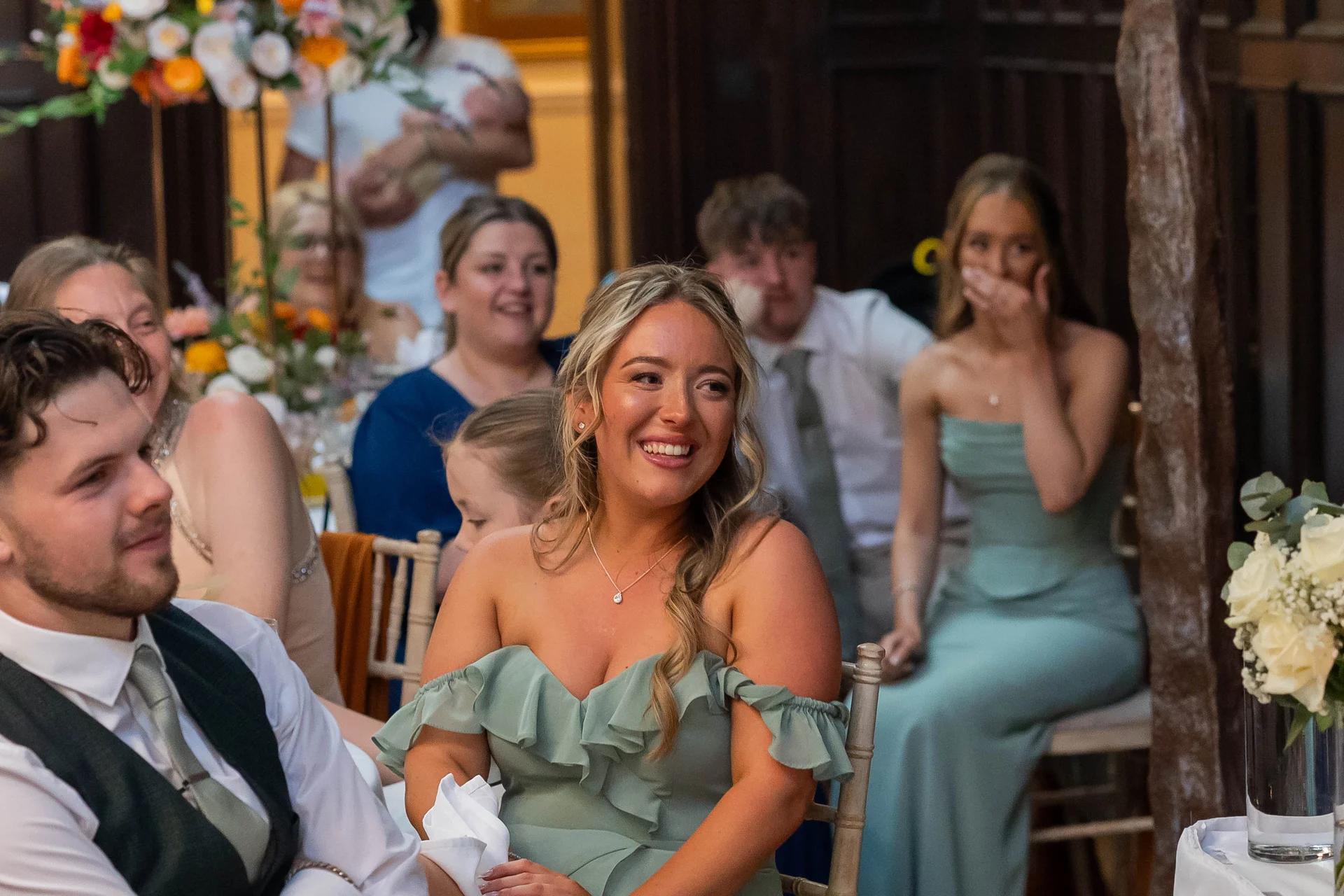 A smiling guest in a sage green off-the-shoulder dress listens happily during the wedding speeches surrounded by family and friends.