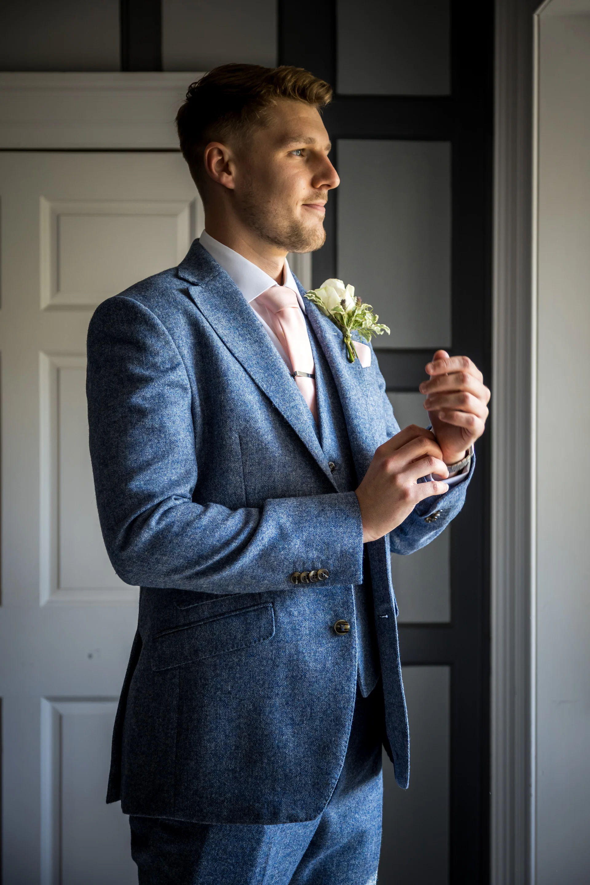 A handsome groom in a blue tweed three piece suit adjusts his cufflinks near a doorway on the morning of his wedding.