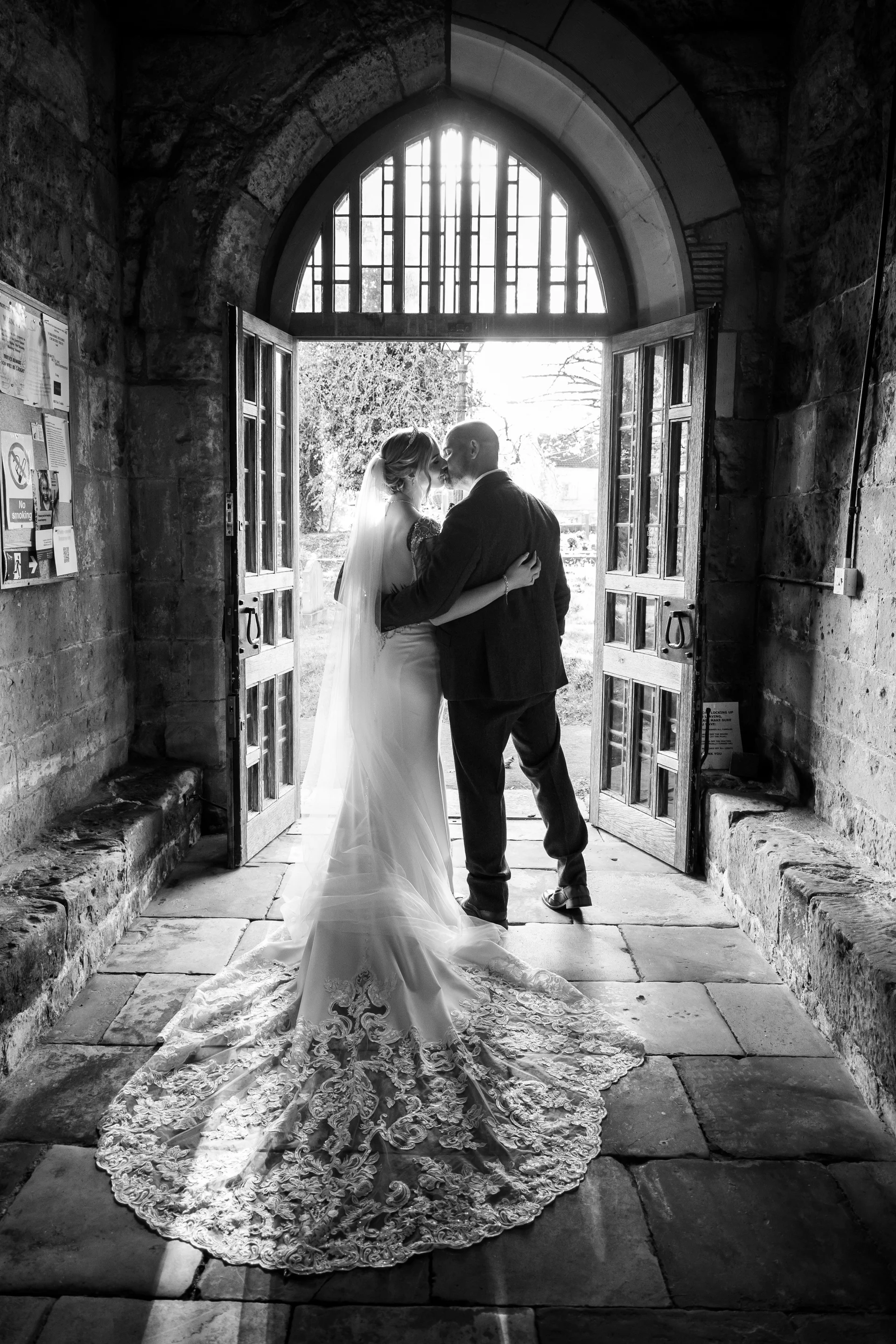 Newlywed bride and groom share a tender kiss framed by the open wooden doors and stone archway of a historic church.