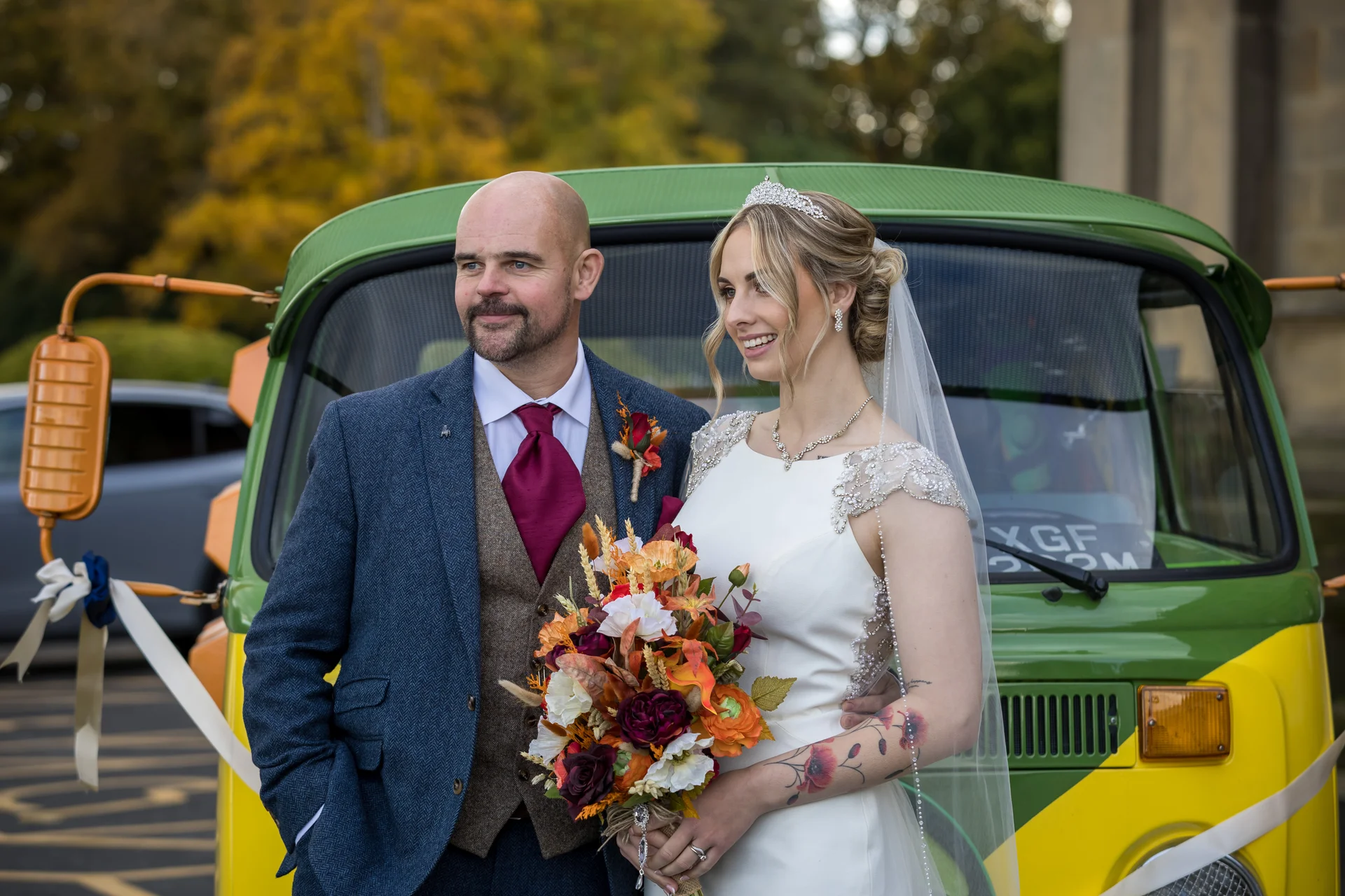A smiling bride with floral tattoos and a crystal tiara poses alongside her groom in a blue tweed suit in front of a vintage green and yellow campervan, holding an autumnal bouquet.