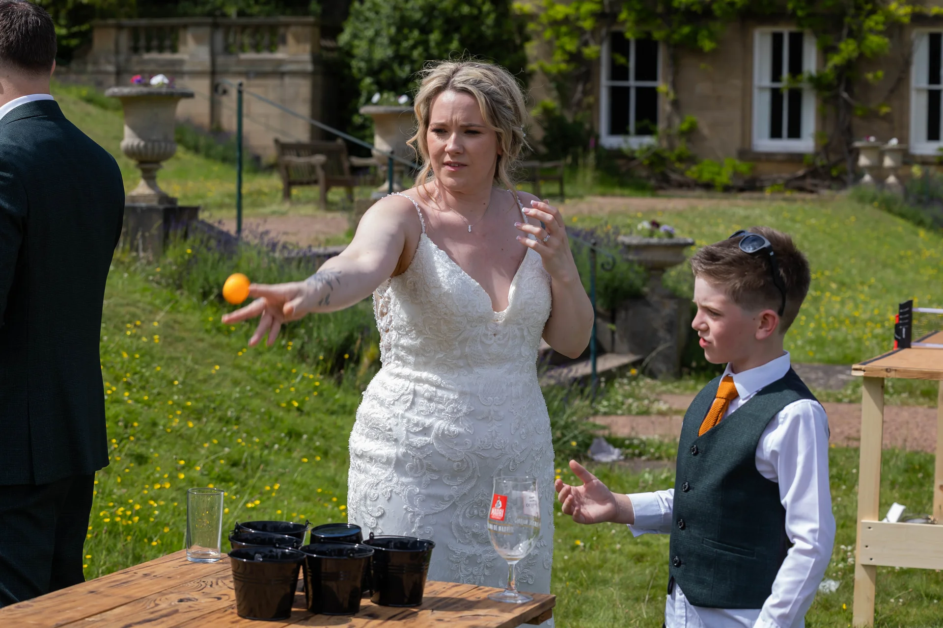 A tattooed bride in a lace wedding dress enthusiastically throws a ball during an outdoor lawn game while a page boy watches on a sunny day.