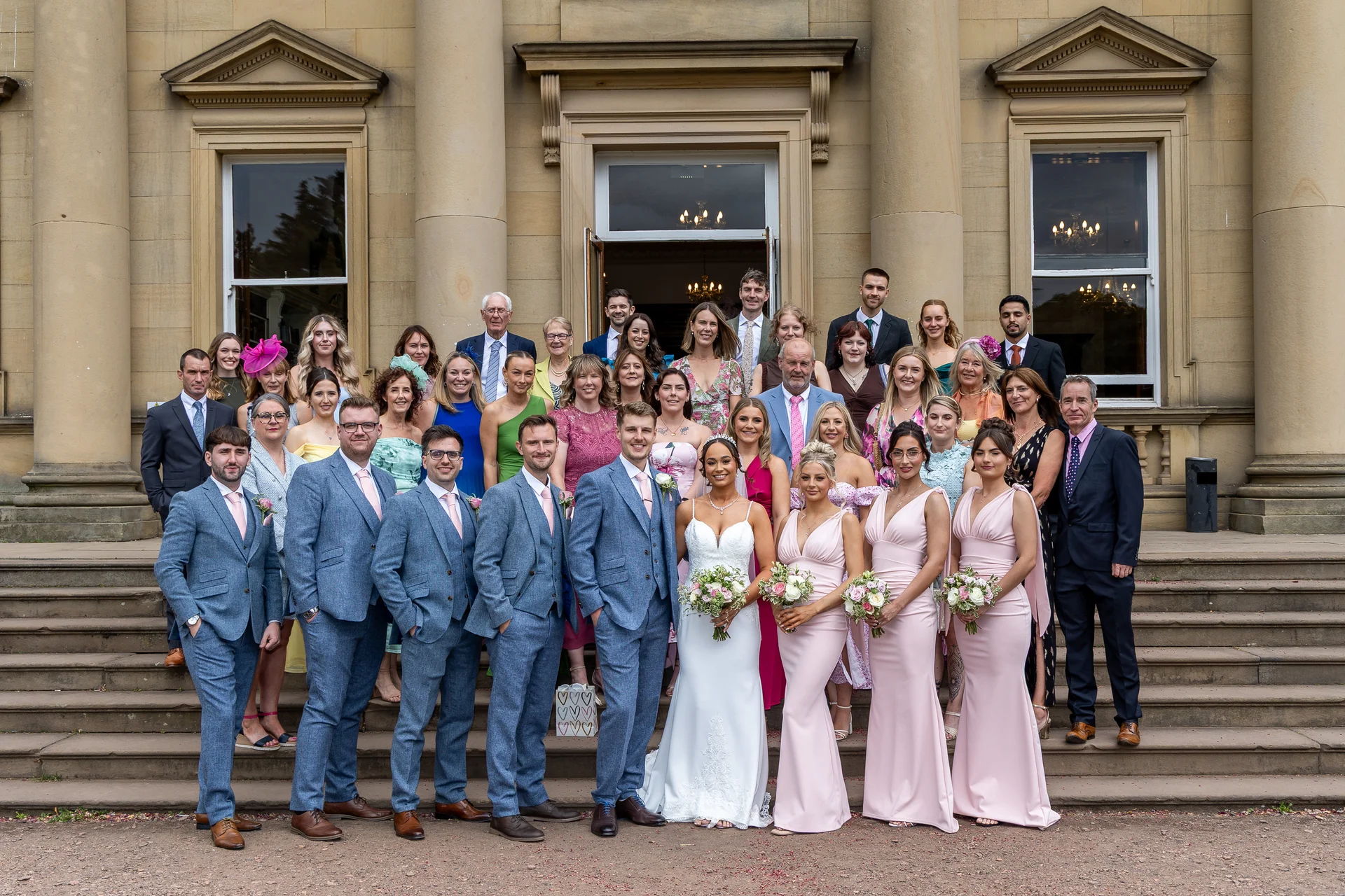 A large wedding party and guests pose for a formal group photograph on the stone steps outside a grand country house.
