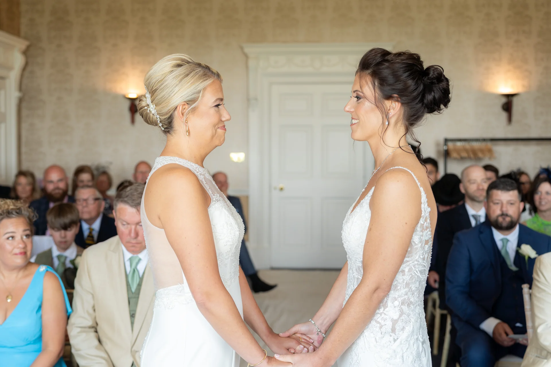 Two brides in white lace wedding dresses stand face-to-face holding hands during their indoor wedding ceremony while seated guests watch on.