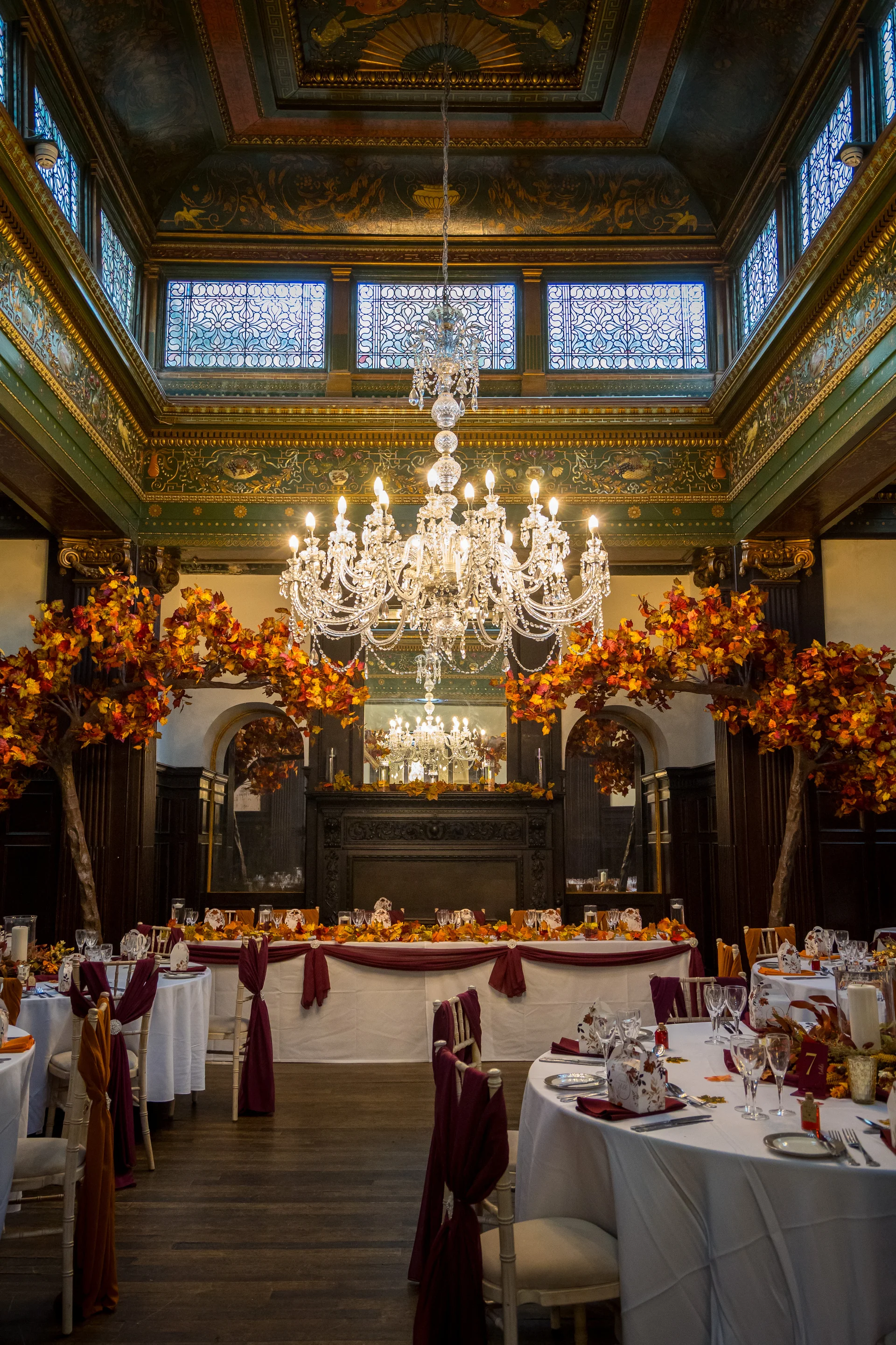 An opulent wedding reception venue features autumn-themed trees flanking a grand top table, illuminated by a magnificent crystal chandelier and surrounded by round tables set with burgundy drapes.