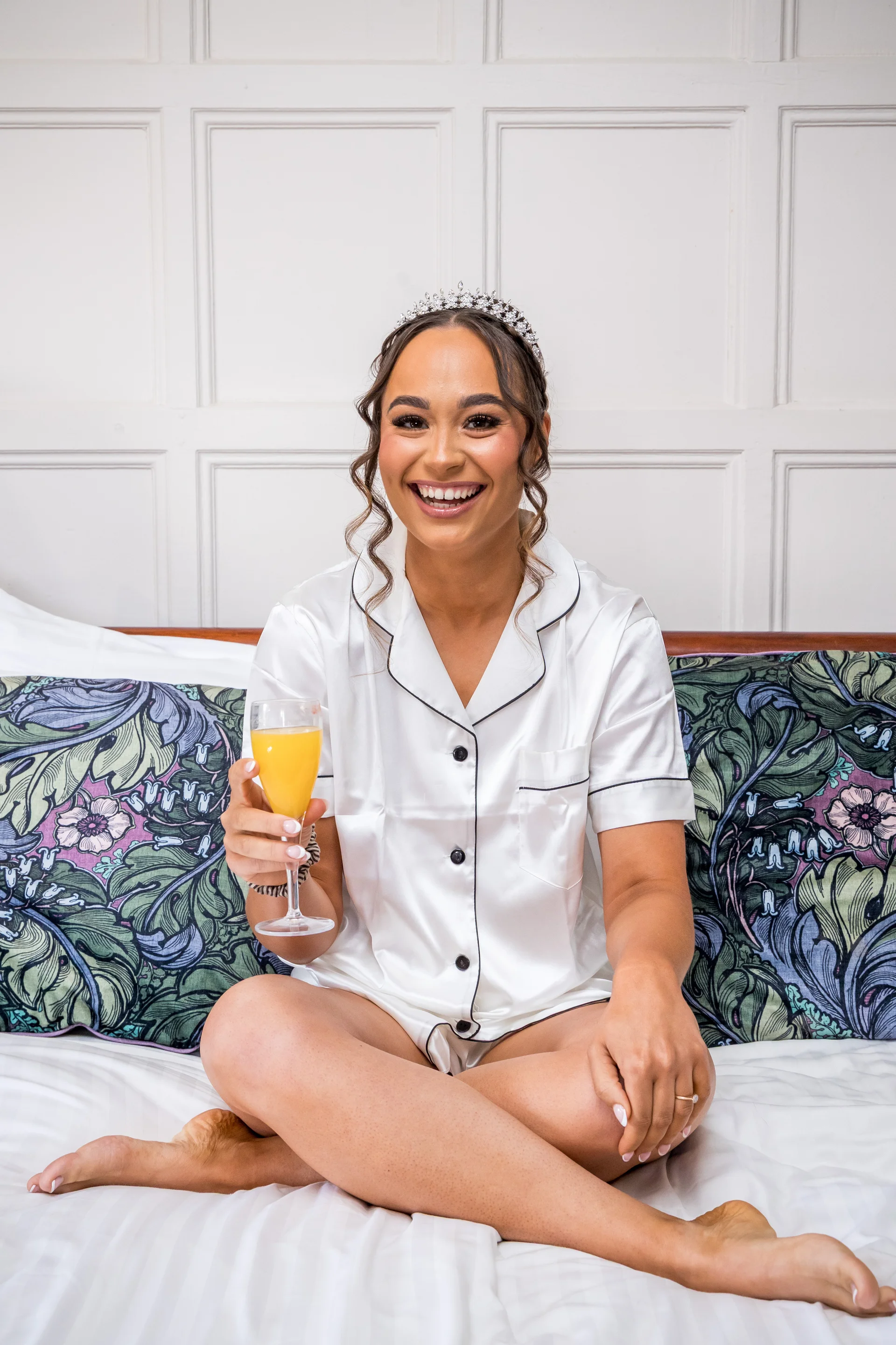 A smiling bride wearing white silk pyjamas and a crystal tiara sits cross-legged on a bed, holding a glass of juice in front of a white panelled wall.