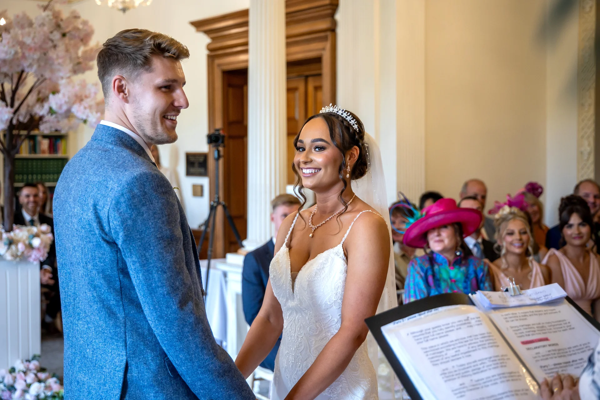 A smiling bride and groom stand together during their civil wedding ceremony, looking at each other happily while surrounded by seated guests.