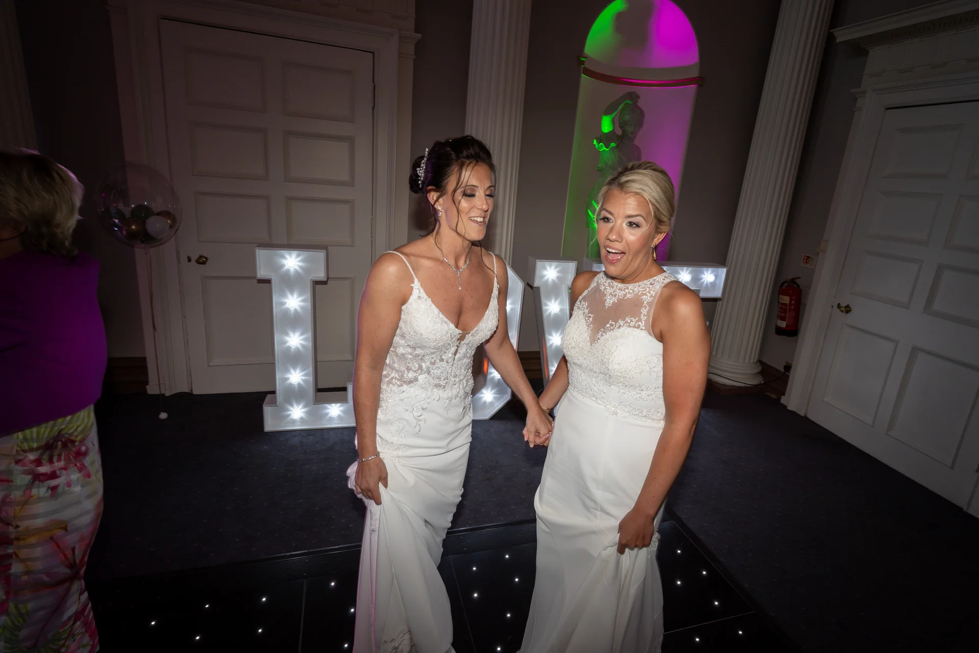 Two smiling brides in white wedding dresses hold hands as they share a joyful moment on the dance floor during their evening reception.