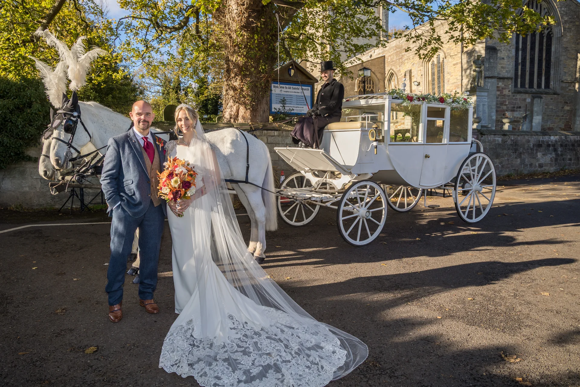 A newlywed couple poses together in front of a historic stone church next to a white horse-drawn carriage on a sunny autumn day.