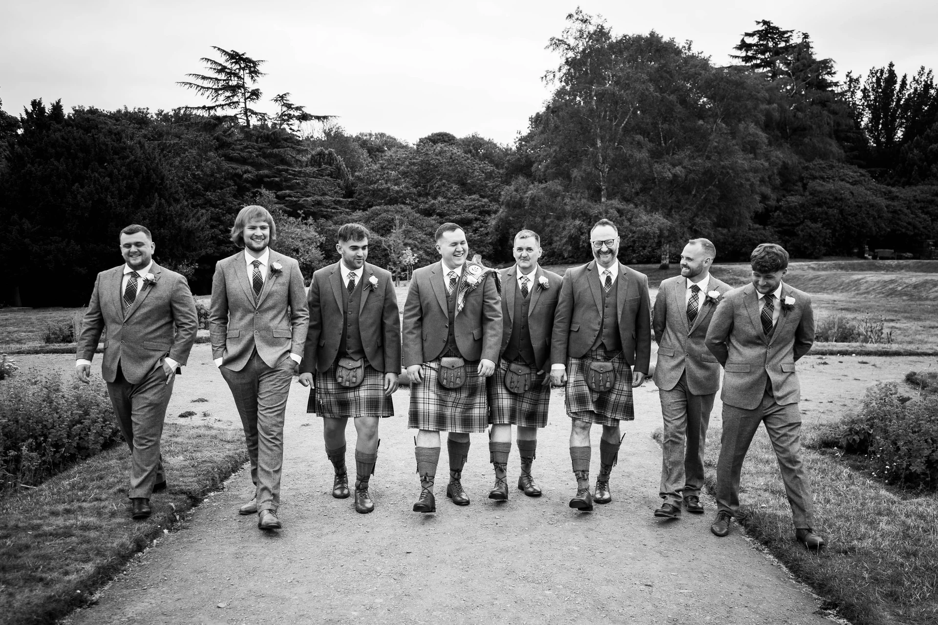 A group of groomsmen wearing tweed suits and tartan kilts walk together down a garden path, smiling and chatting.