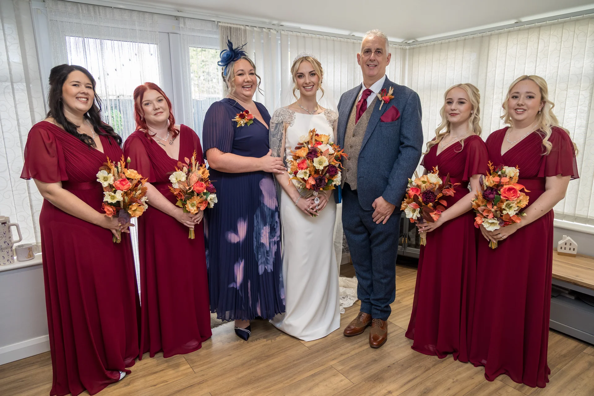 A bridal party poses indoors featuring bridesmaids in burgundy dresses, a mother of the bride in navy, and the bride and groom holding autumnal bouquets.