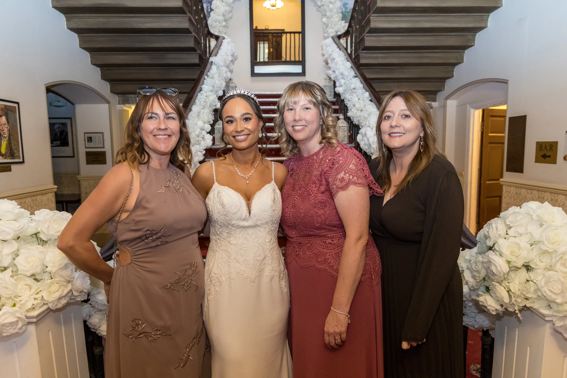 A smiling bride in a white lace a-line gown stands alongside three female guests in front of a grand staircase decorated with white floral garlands.