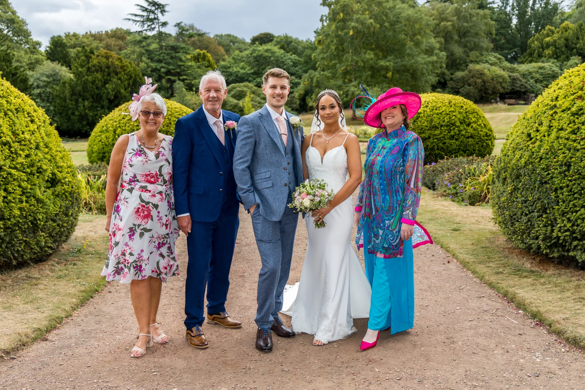 A newlywed couple and family members pose together on a garden path flanked by neatly trimmed bushes on a bright summer day.