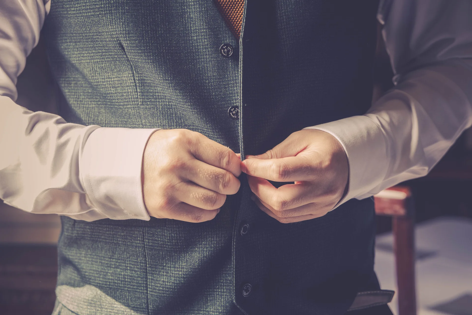 A groom is shown from the torso up buttoning up his textured grey-green tweed wedding waistcoat over a crisp white shirt.