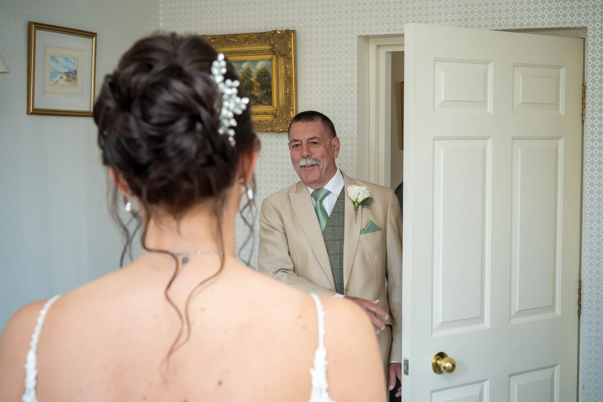A father smiles warmly upon seeing his daughter for the first time in her wedding dress, captured from over her shoulder as he enters the room.