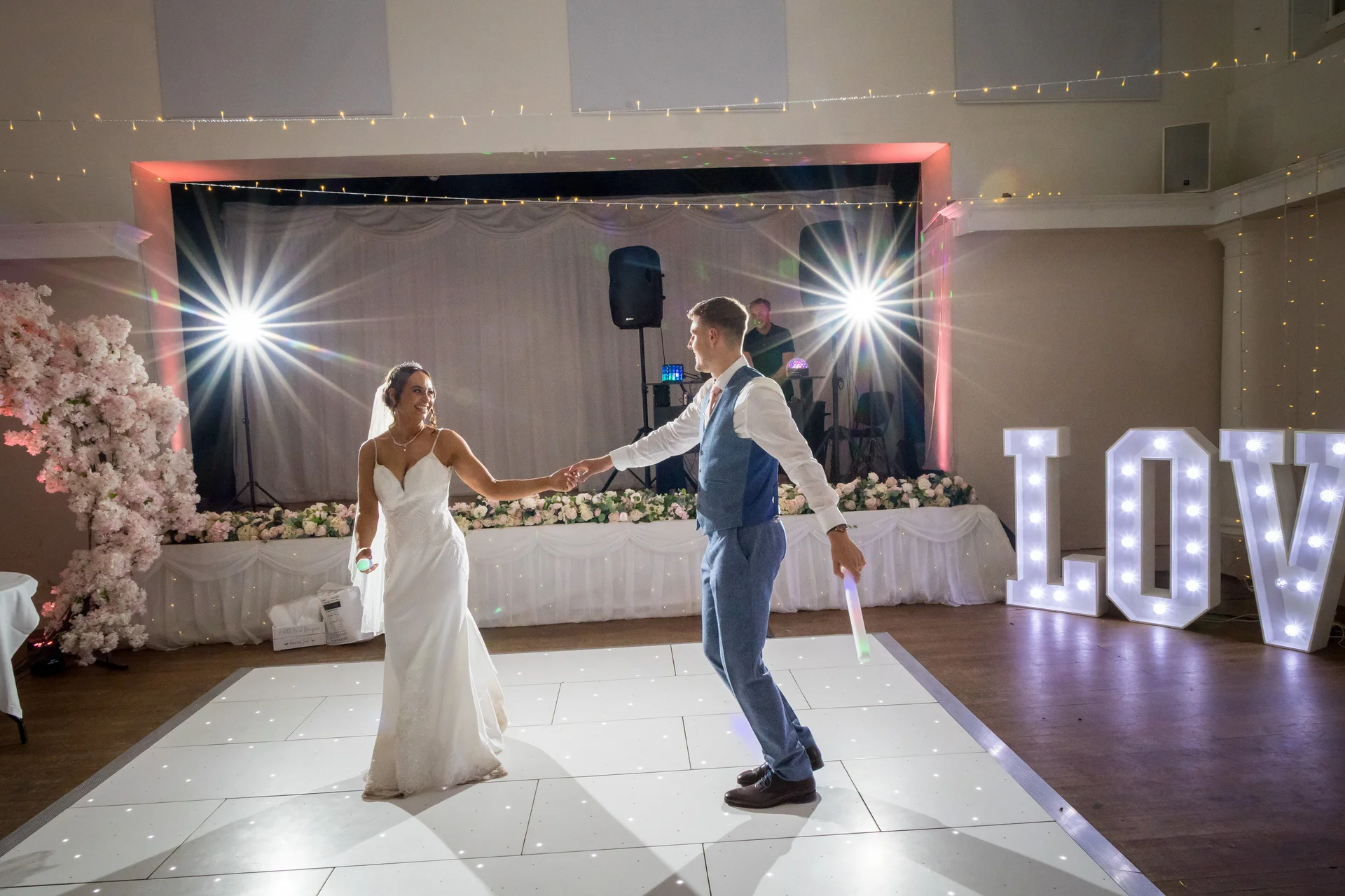 Newlyweds share their first dance on an illuminated LED dance floor, holding glowing batons while large illuminated love letters glow nearby.