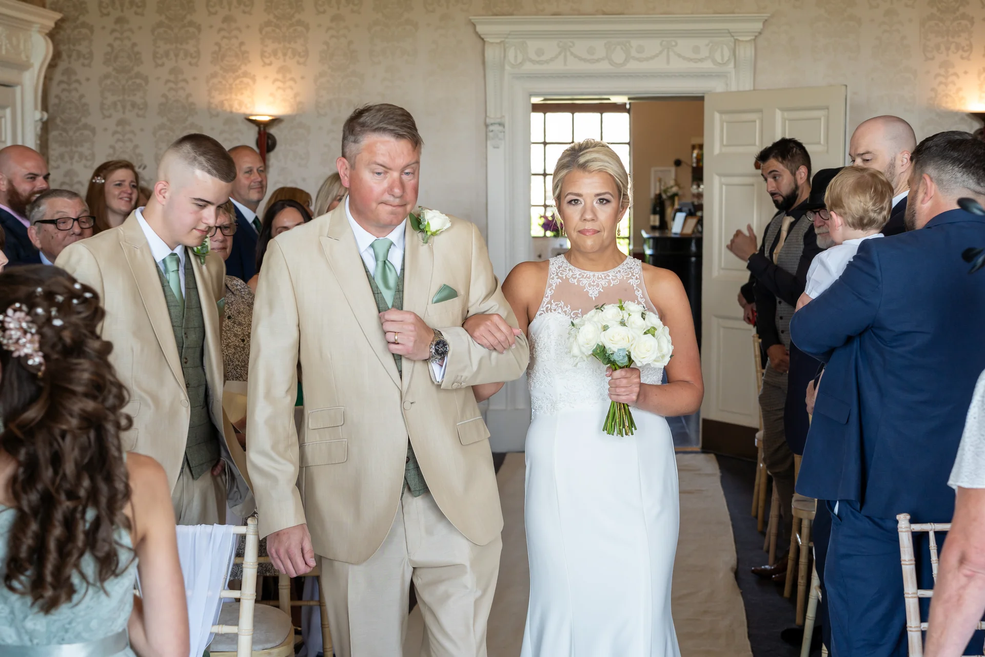 A bride walks down the aisle arm in arm with her father, holding a bouquet of white roses during an indoor wedding ceremony.