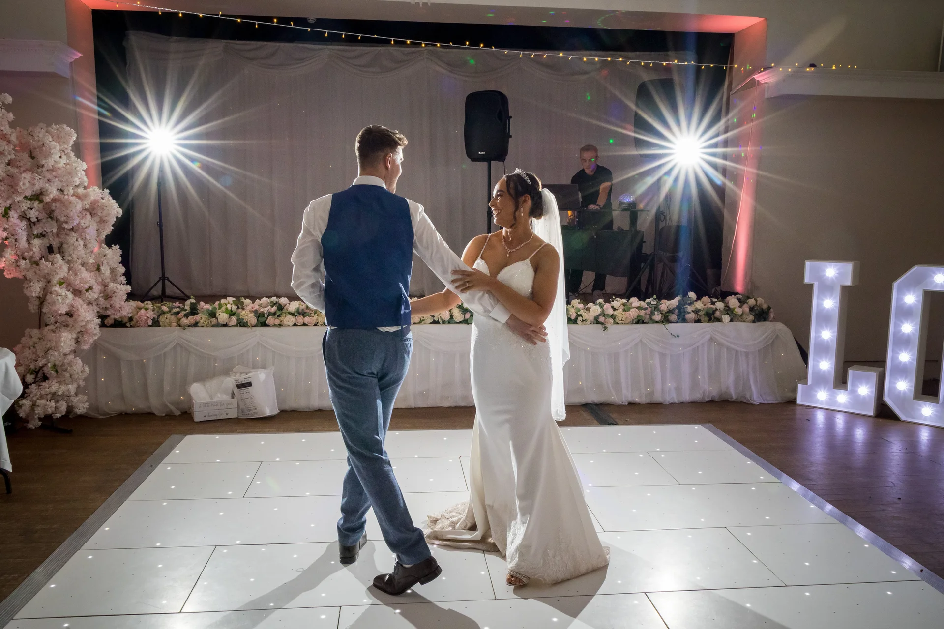 Newlyweds share their first dance on a lighted white dance floor under stage spotlights, with a floral backdrop and glowing marquee letters.