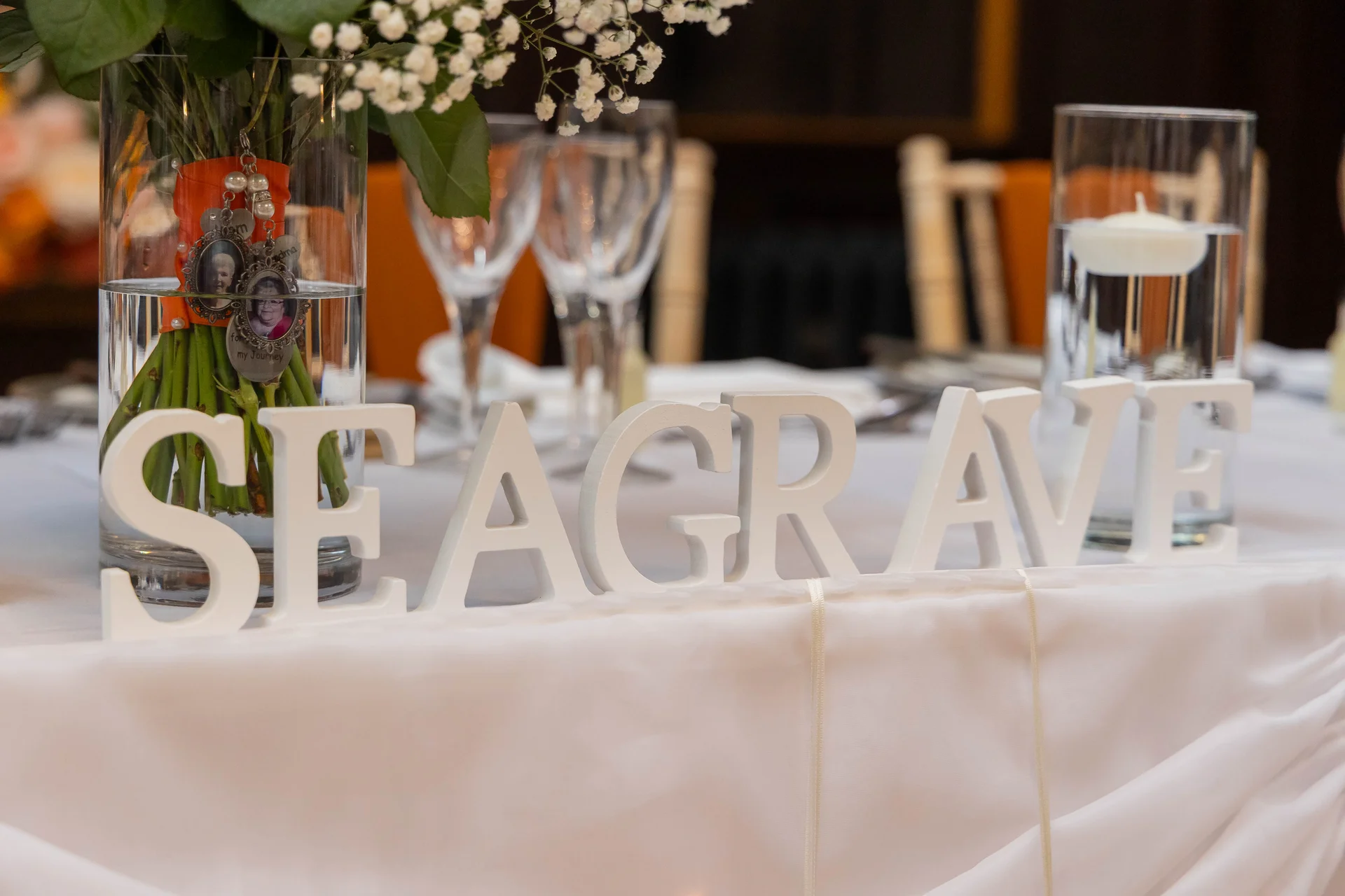 A close-up shot of a wedding top table featuring white freestanding letters spelling a family name, accompanied by a tall floral arrangement with memorial charms and a floating candle in a glass cylinder.