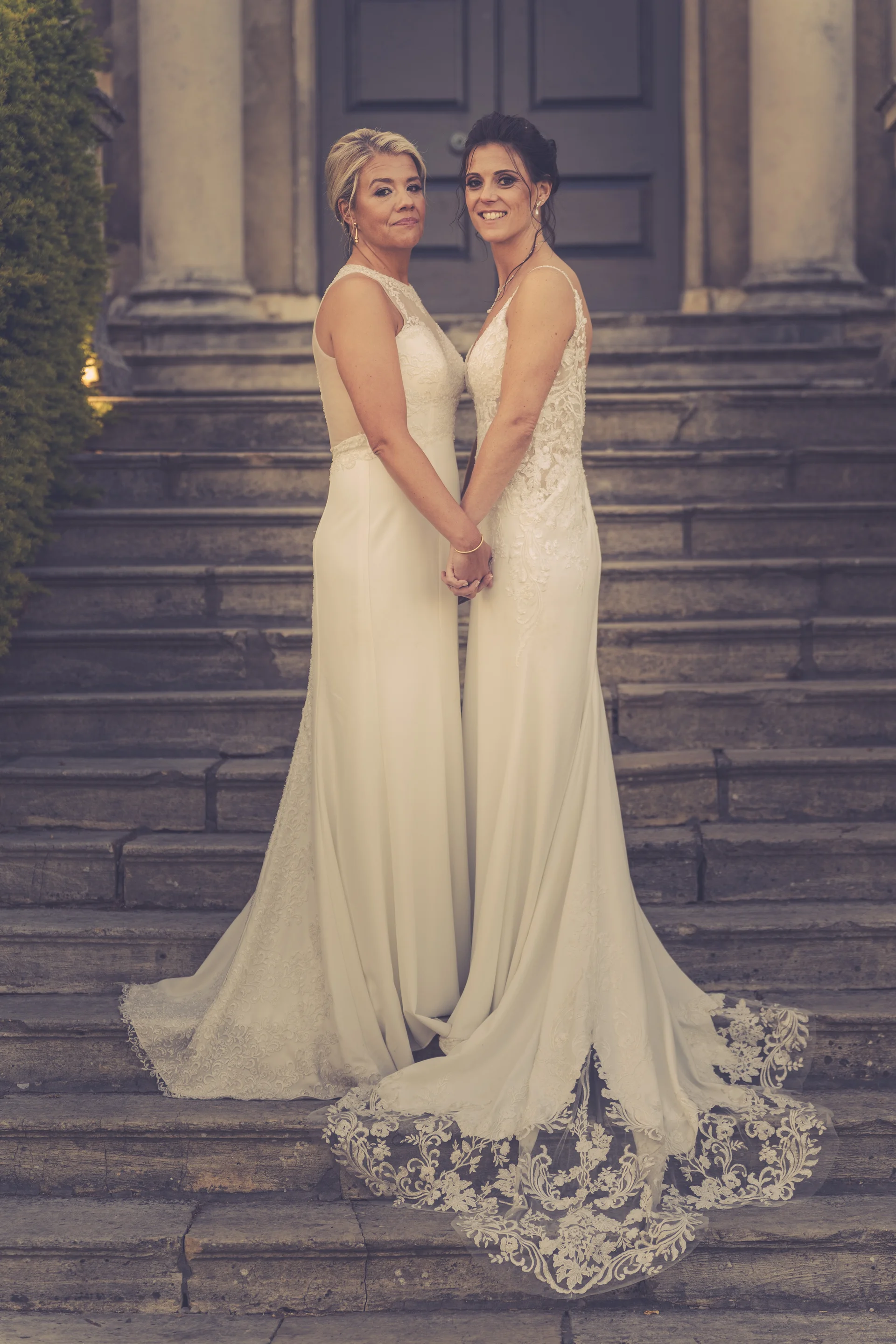 Two smiling brides holding hands while standing together on grand stone steps outside a historic venue.