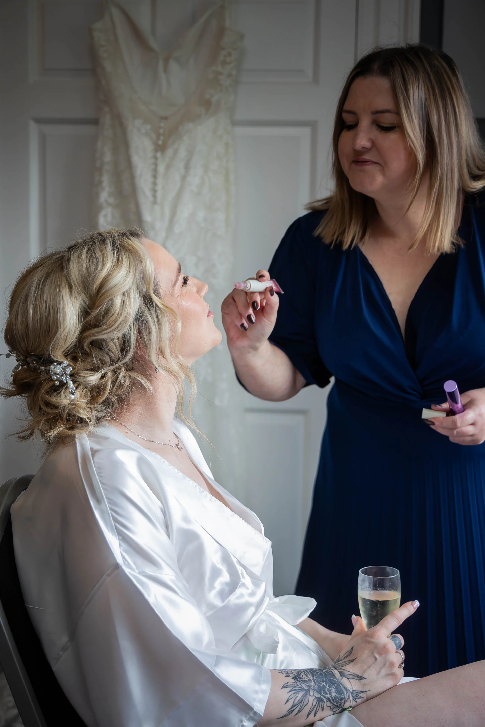A blonde bride wearing a white satin robe and half-up hairstyle sits holding a glass of champagne while having her makeup applied.