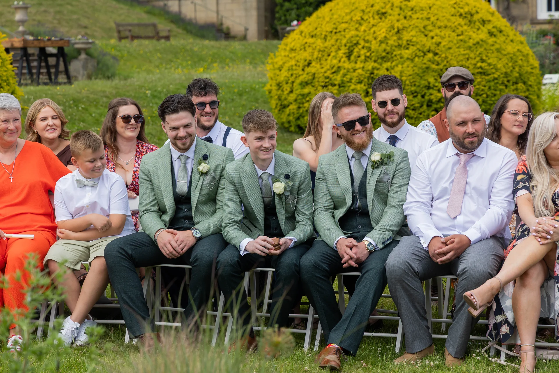 A group of smiling groomsmen and wedding guests seated outdoors on chairs during a sunny summer ceremony.