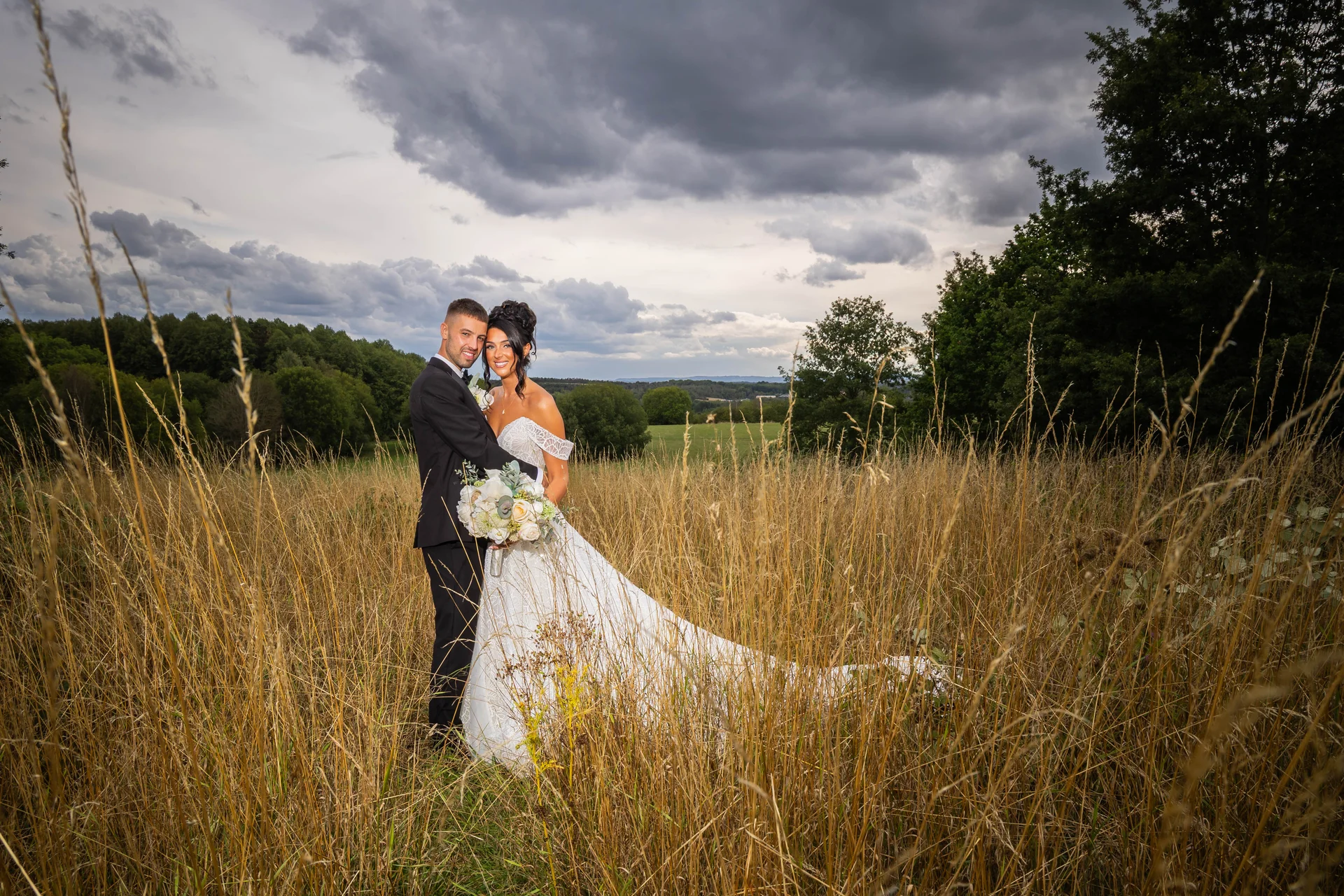 A bride and groom pose together in a tall grass field against a dramatic, cloudy sky backdrop.