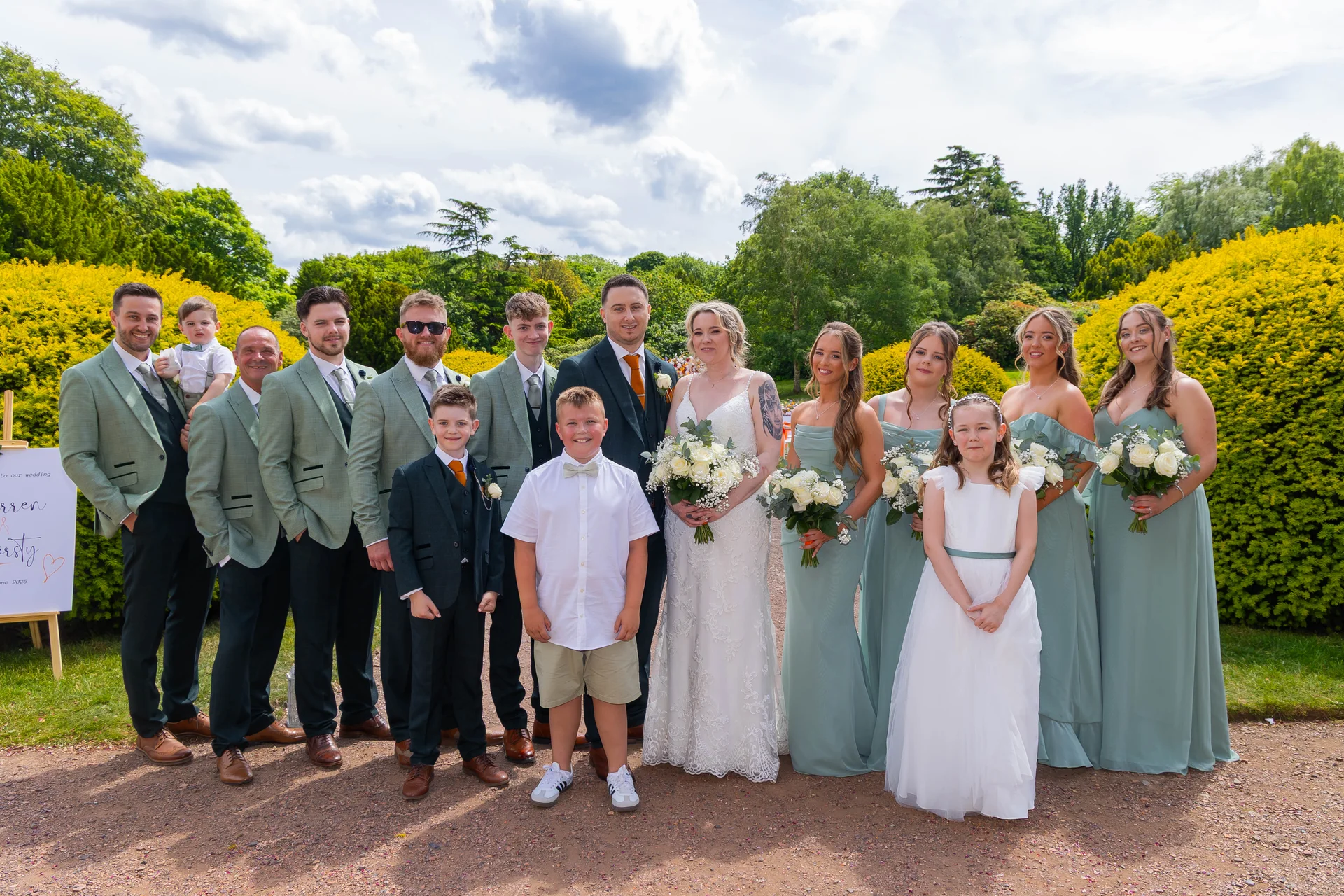 A full bridal party poses outdoors in a sunlit garden, featuring the bride in a white lace gown, groom in a dark suit, groomsmen in sage green suits, and bridesmaids in matching sage dresses.