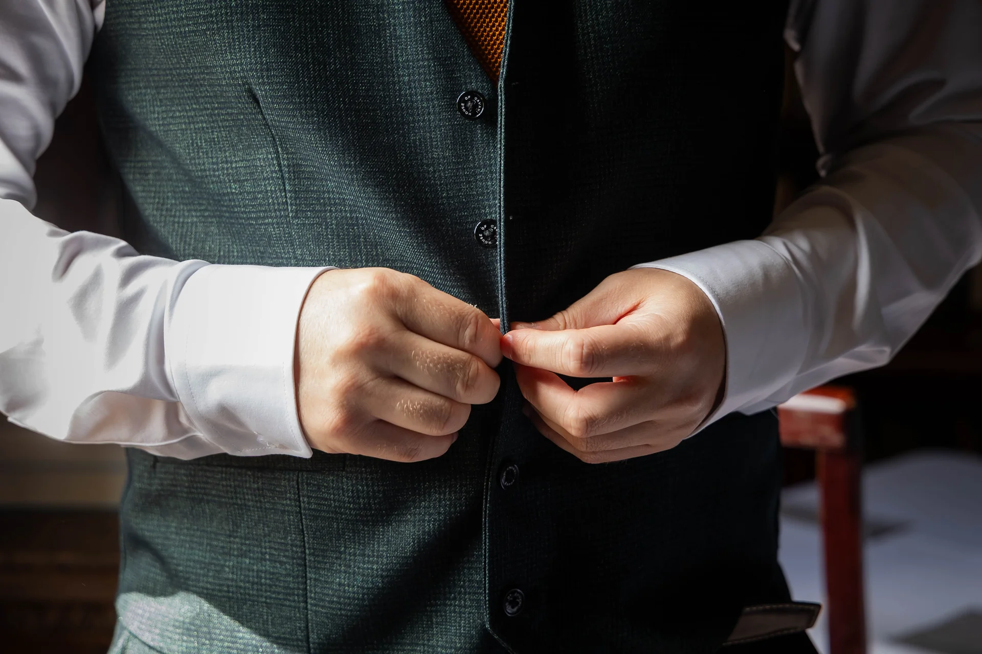 A groom carefully buttoning up his green textured waistcoat during morning wedding preparations.