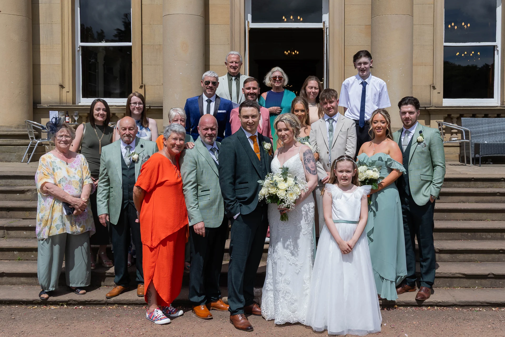 A large wedding party and family members pose for a formal group photograph on the stone steps outside a grand country house venue.