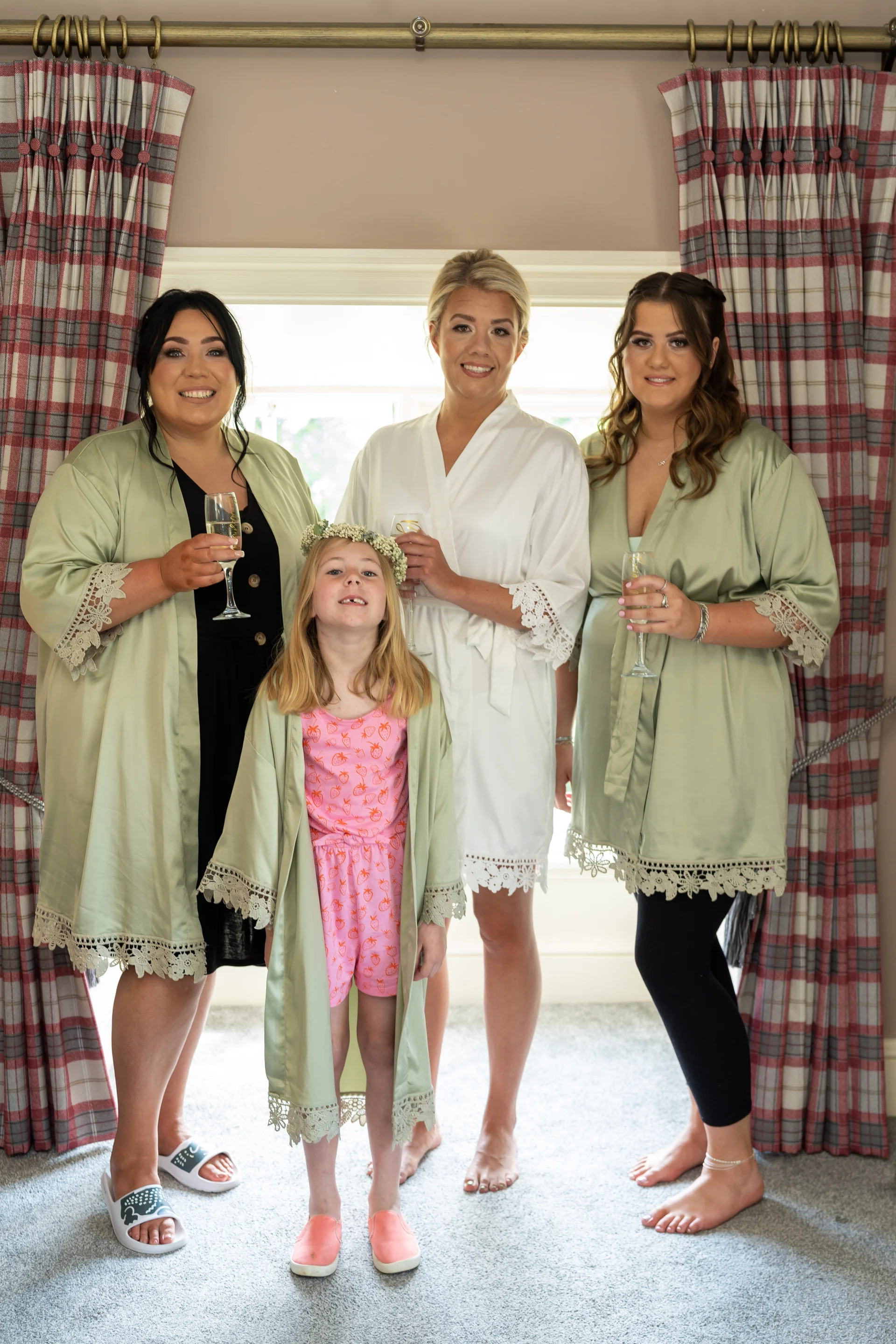 A smiling bride in a white robe stands with two bridesmaids in sage robes and a young flower girl holding champagne glasses indoors.